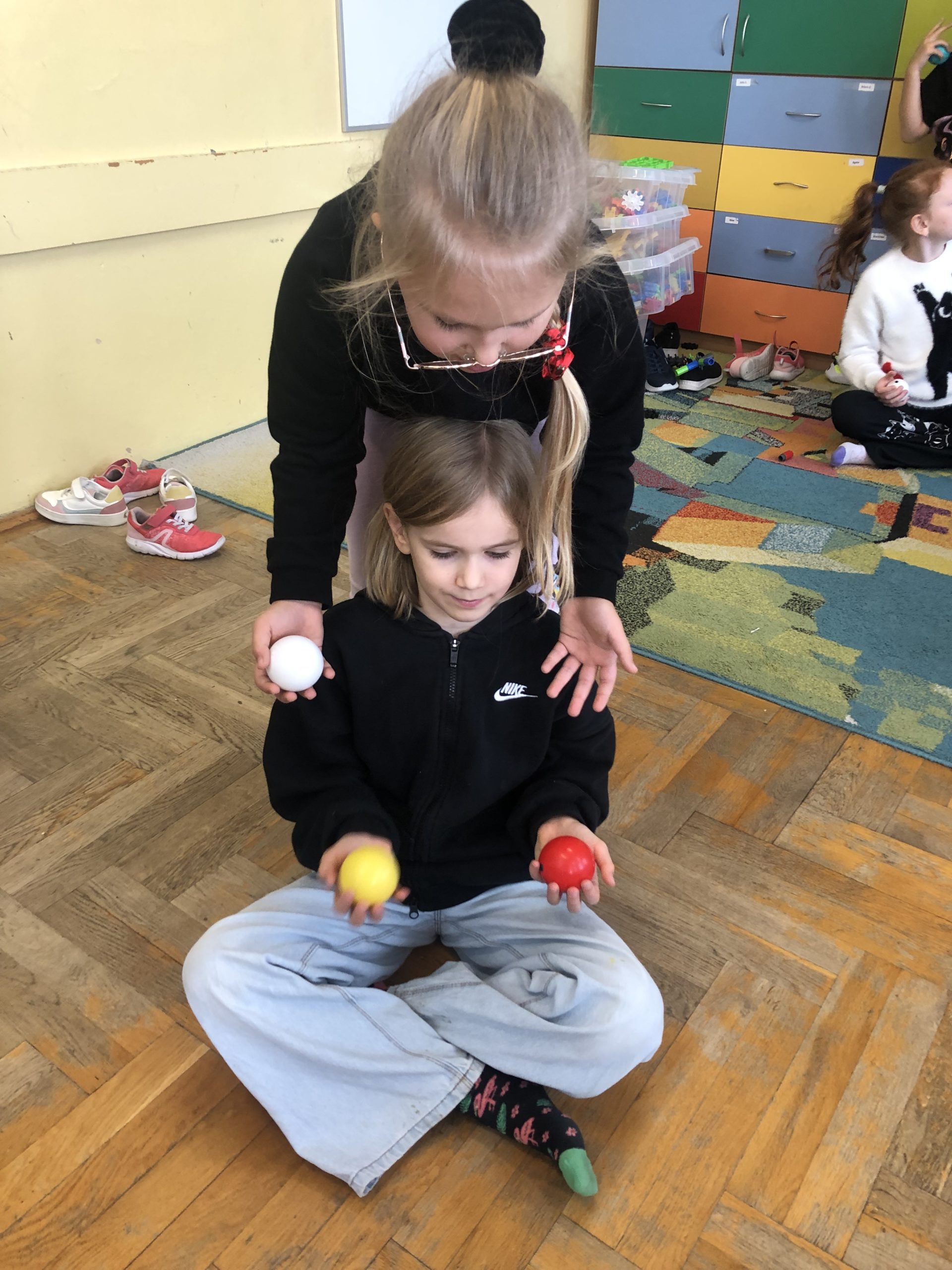 Dwie dziewczynki, jedna siedzi na podłodze, druga nachyla się nad nią. W rękach trzymają piłeczki cyrkowe.