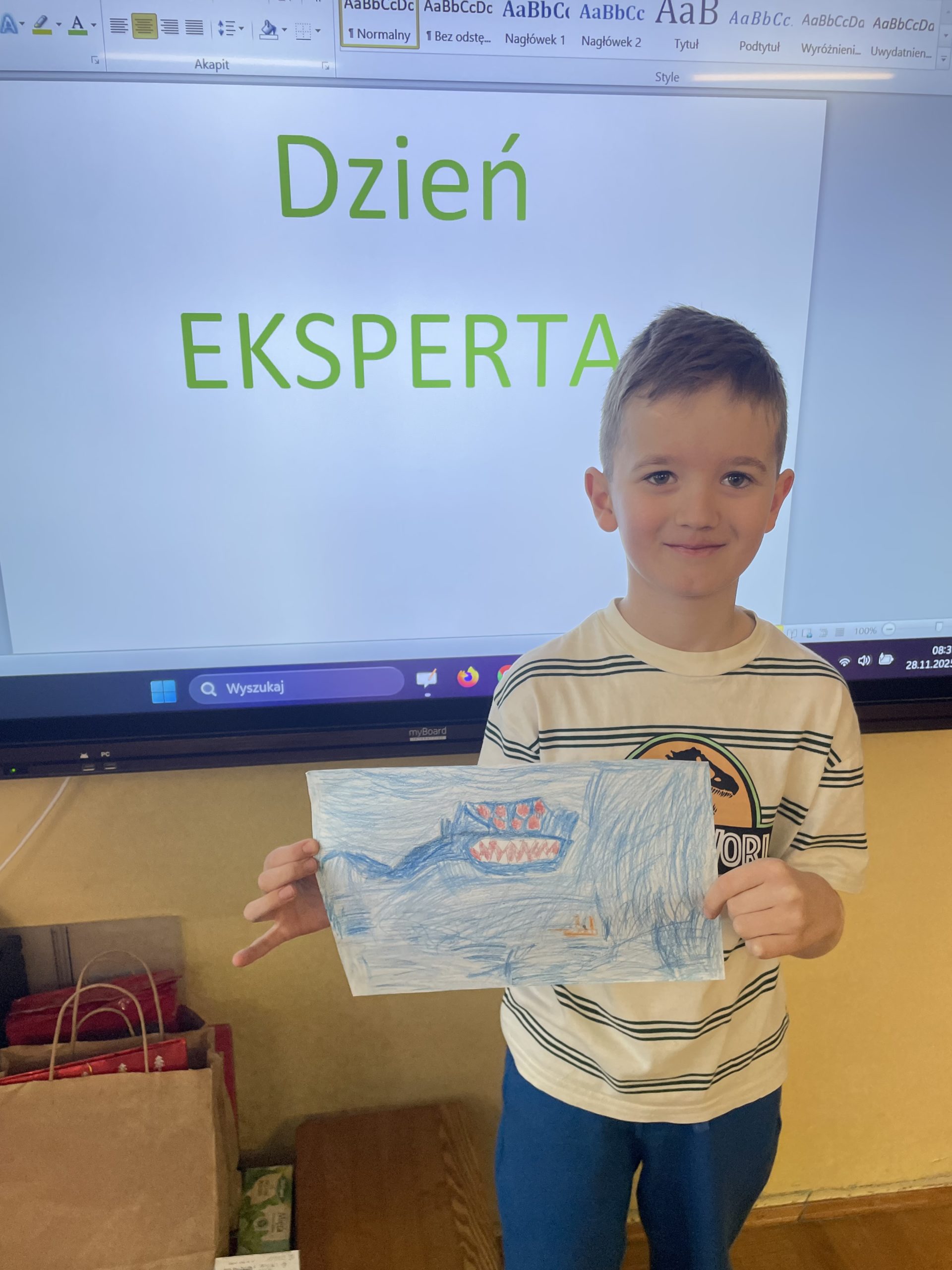 Chłopiec stoi przed tablicą z napisem Dzień Eksperta, w rękach trzyma rysunek morskiego stwora.