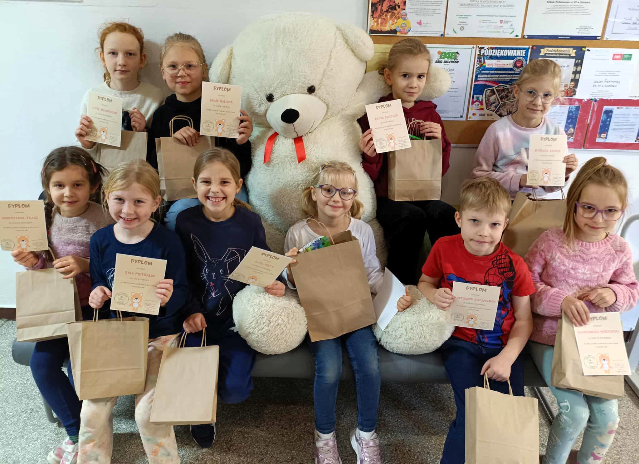 Grupa jedenastu uczniów siedzi na ławce obok dużego, białego pluszowego misia. Dzieci uśmiechają się i trzymają dyplomy oraz papierowe torebki z nagrodami.