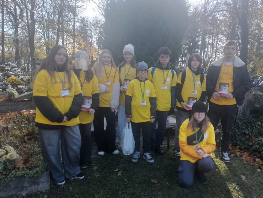 Grupa wolontariuszy w koszulkach stoi na cmentarzu. Na szyi mają identyfikatory. Trzy osoby trzymają w ręku skarbonkę.