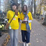 Dwie dziewczyny w koszulkach wolontariuszy i identyfikatorami na szyi stoją obok siebie na cmentarzu. W ręku jedna z nich ma skarbonkę. Ubiór i aura wskazują na jesienną pogodę.