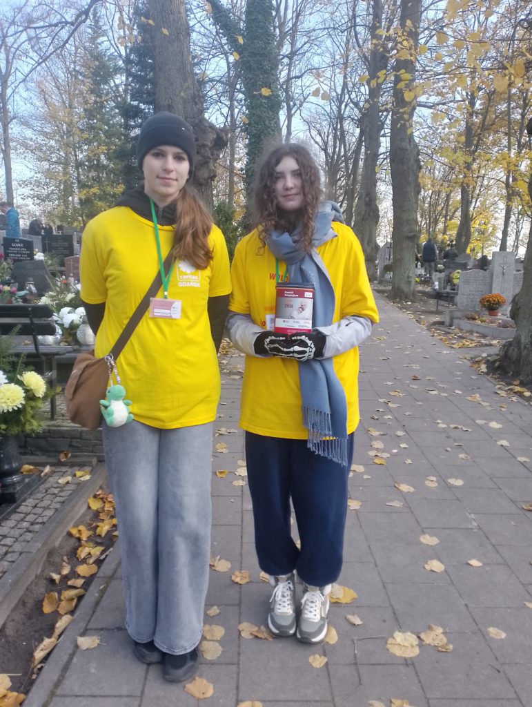 Dwie dziewczyny w koszulkach wolontariuszy i identyfikatorami na szyi stoją obok siebie na cmentarzu. W ręku jedna z nich ma skarbonkę. Ubiór i aura wskazują na jesienną pogodę.