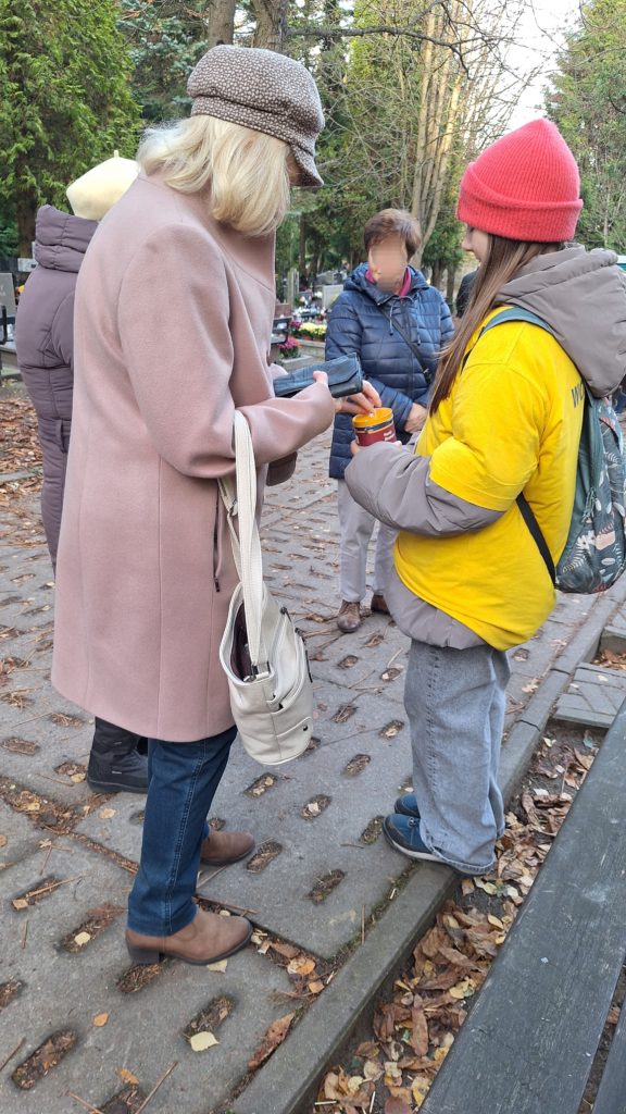 Trzy starsze kobiety stoją przy dziewczynce w koszulce wolontariusza, która w ręku trzyma skarbonkę. Jedna z nich wrzuca do puszki pieniądze.