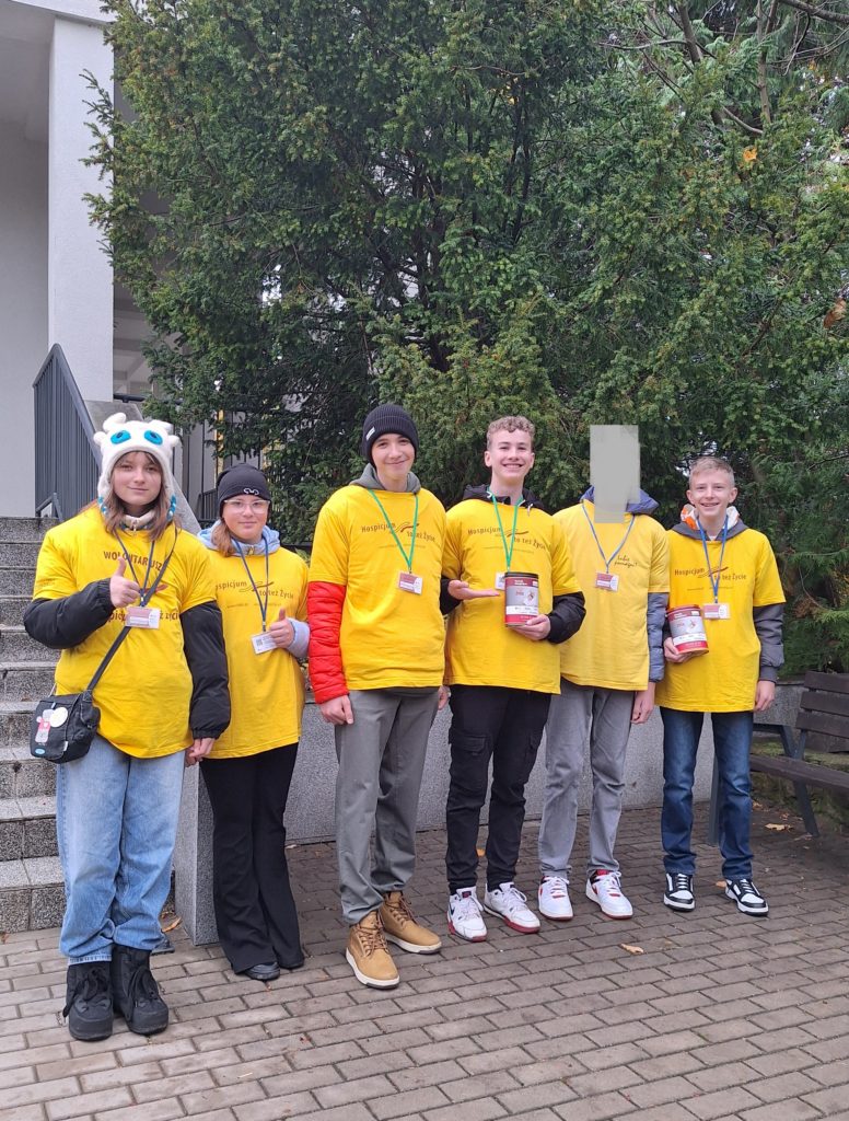 Sześcioro wolontariuszy stoi obok siebie. Ubrani są w koszulki wolontariuszy, na szyi mają identyfikatory. Dwóch chłopców trzyma w ręku skarbonki, a dwie dziewczynki pokazują kciuki do góry.