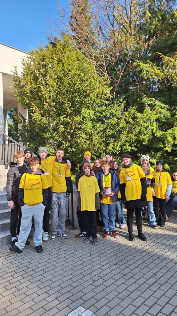 Grupa wolontariuszy w koszulkach stoi na cmentarzu. Na szyi mają identyfikatory. Dwie osoby trzymają w ręku skarbonkę. Świeci słońce. Jest radosna atmosfera.