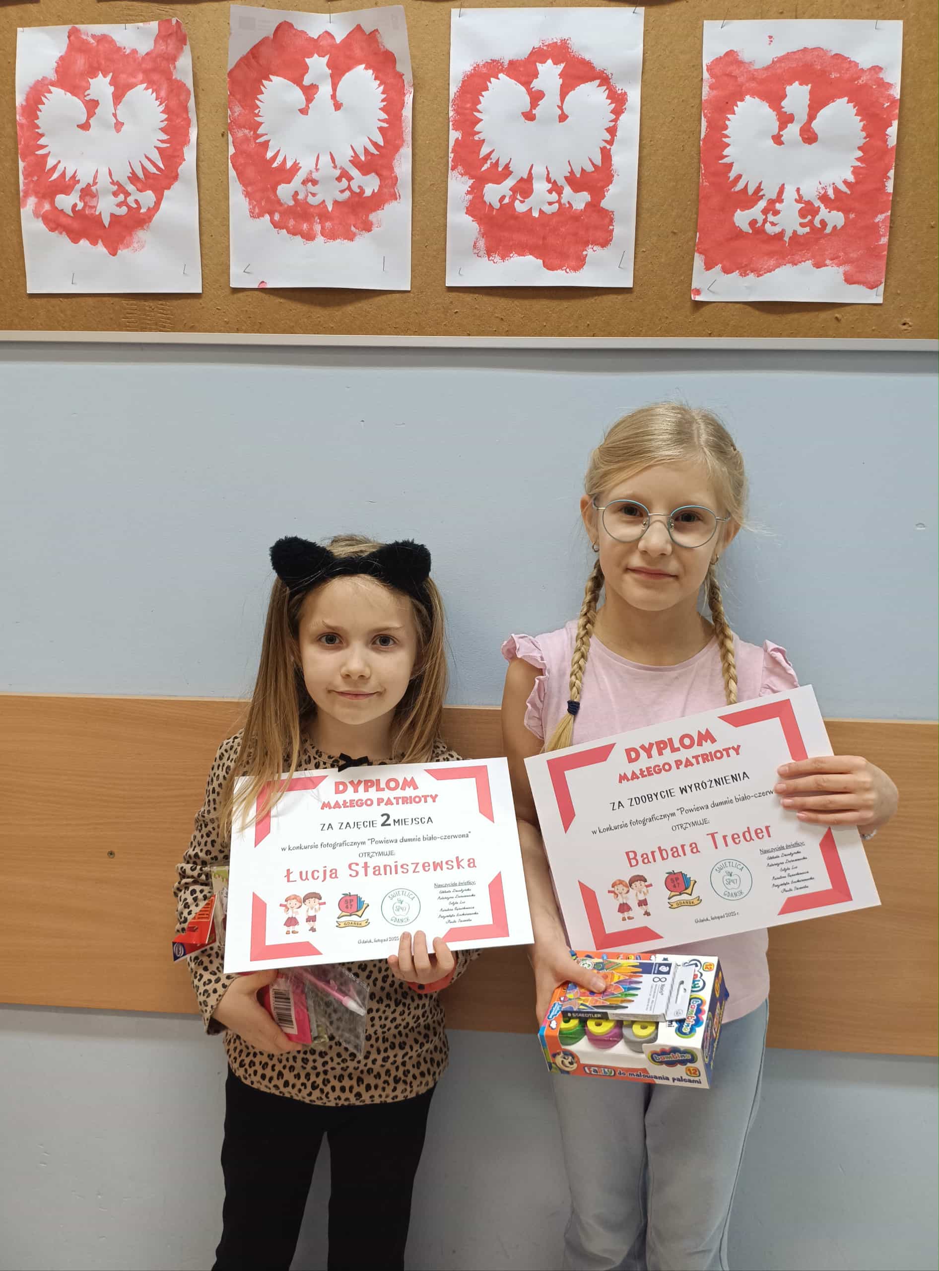 Dwie dziewczynki stoją obok siebie i trzymają dyplomy za udział w świetlicowym konkursie patriotycznym. W tle na tablicy znajdują się prace przedstawiające biało-czerwone godło namalowane farbami.