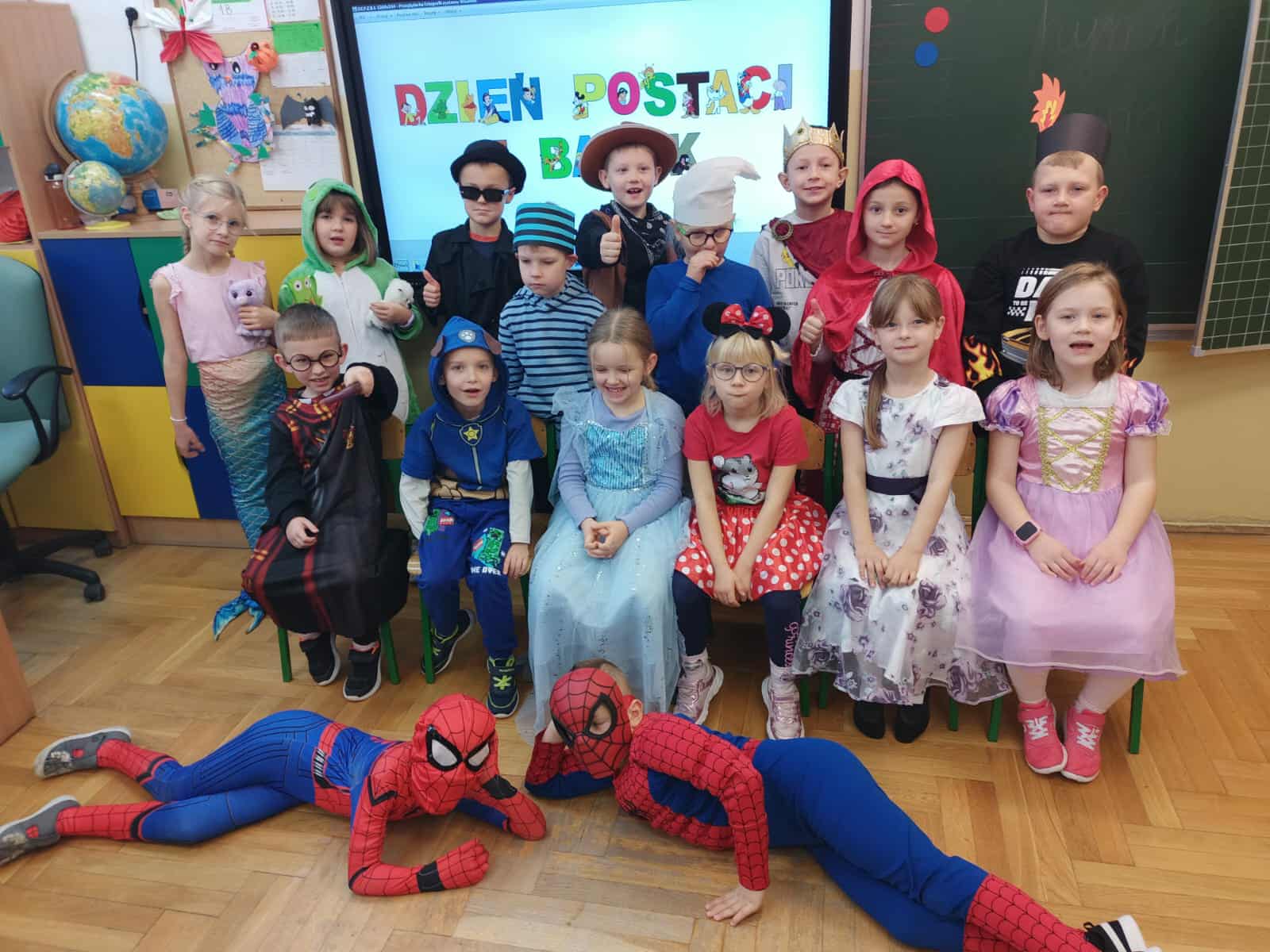 Uczniowie klasy 1a przebrani za postacie z bajek – m.in. królewny, wróżki, superbohaterowie i bajkowe zwierzątka – pozują wspólnie w sali podczas Dnia Postaci z Bajek.