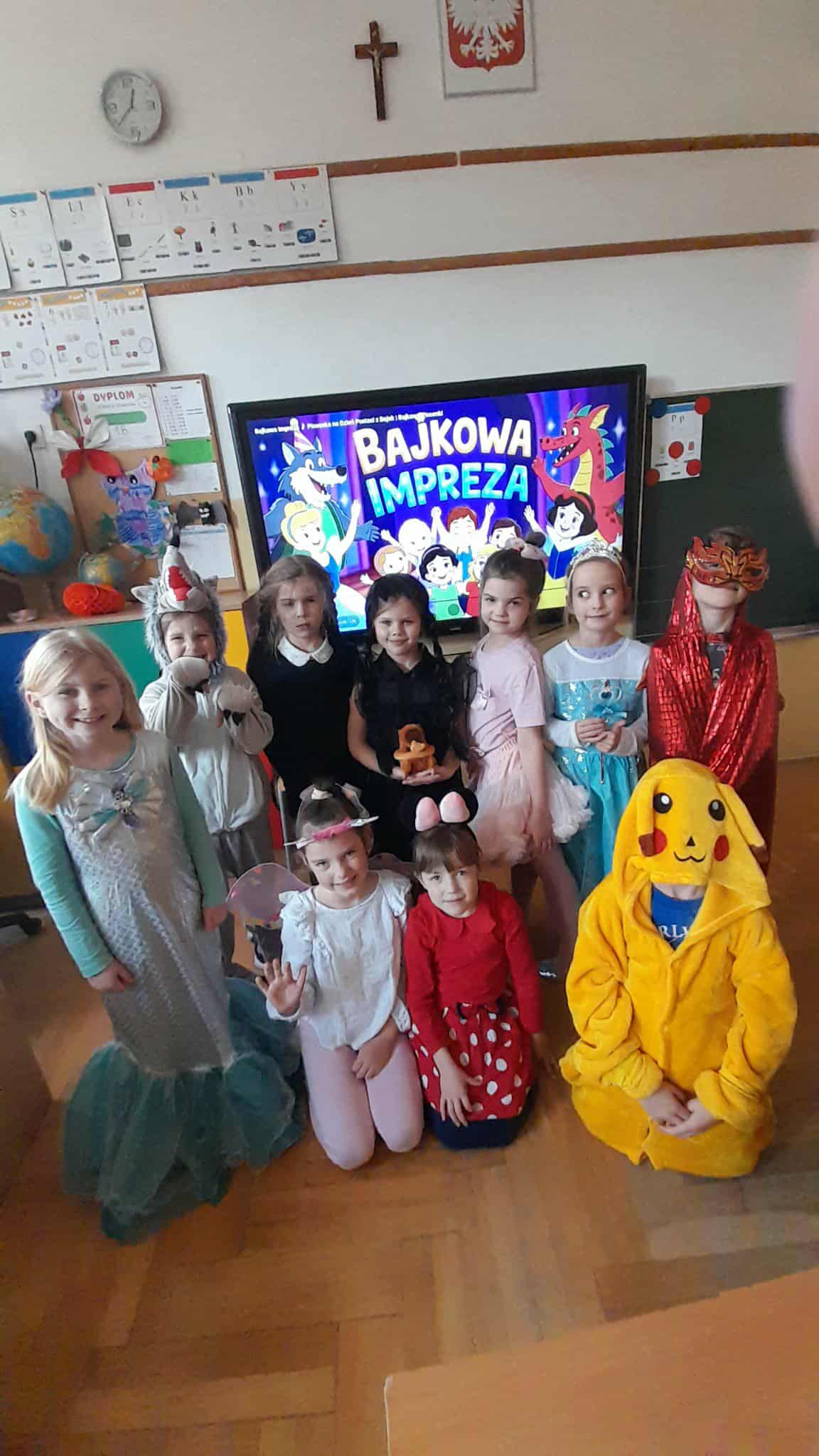 Grupa dzieci z klasy 1b w kolorowych przebraniach bajkowych postaci pozuje w sali lekcyjnej. W tle ekran z napisem „Bajkowa impreza”.