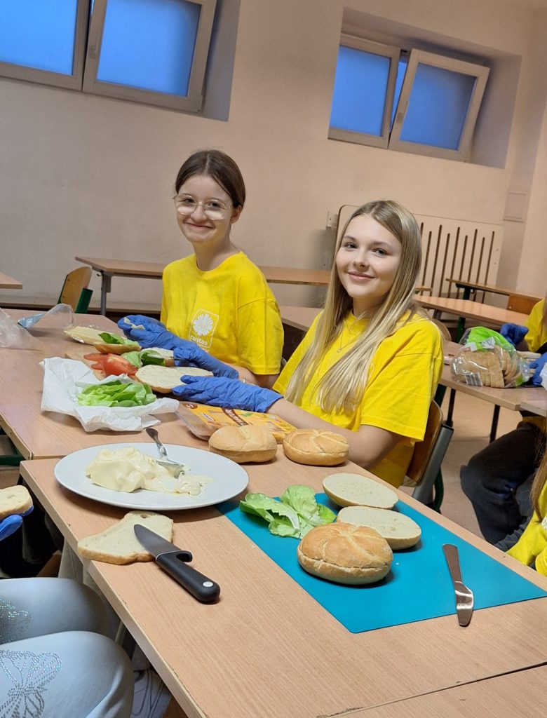 Dwie dziewczynki w koszulkach wolontariuszy siedzą przy stole. Na dłoniach mają rękawiczki jednorazowe i robią kanapki. Na stole leżą bułki, warzywa i inne produkty.