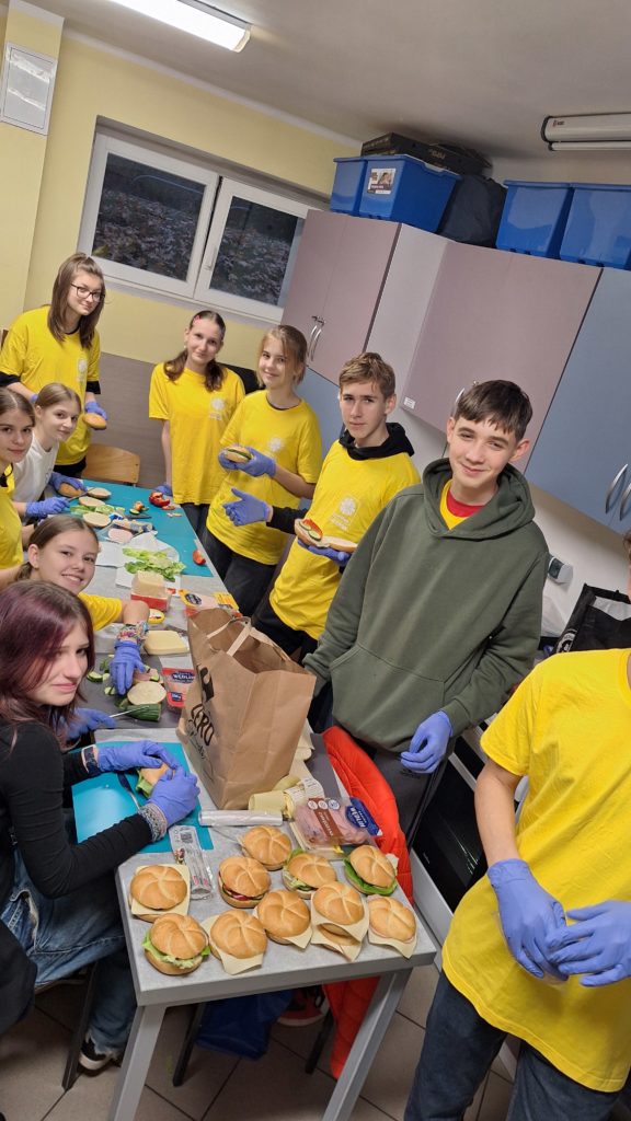Grupa młodzieży w koszulkach wolontariuszy wokół stołu produkują kanapki w szkolnej sali. Na stole widoczne są świeże produkty spożywcze oraz gotowe kanapki.