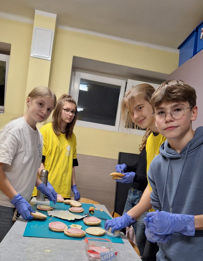 Trzy dziewczyny i chłopak stoją przy stole, na którym leżą rozkrojone bułki. Trwa produkcja kanapek. Na dłoniach mają rękawiczki jednorazowe.