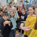 Grupa uczniów stoi z uniesionymi rękoma do góry, w których trzymają kanapki. Przed nimi stoi dziewczyna w koszulce wolontariusza z tacą w ręku, na której znajdują się kanapki.