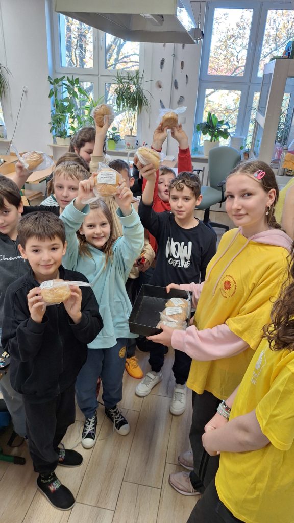 Grupa uczniów stoi z uniesionymi rękoma do góry, w których trzymają kanapki. Przed nimi stoi dziewczyna w koszulce wolontariusza z tacą w ręku, na której znajdują się kanapki.