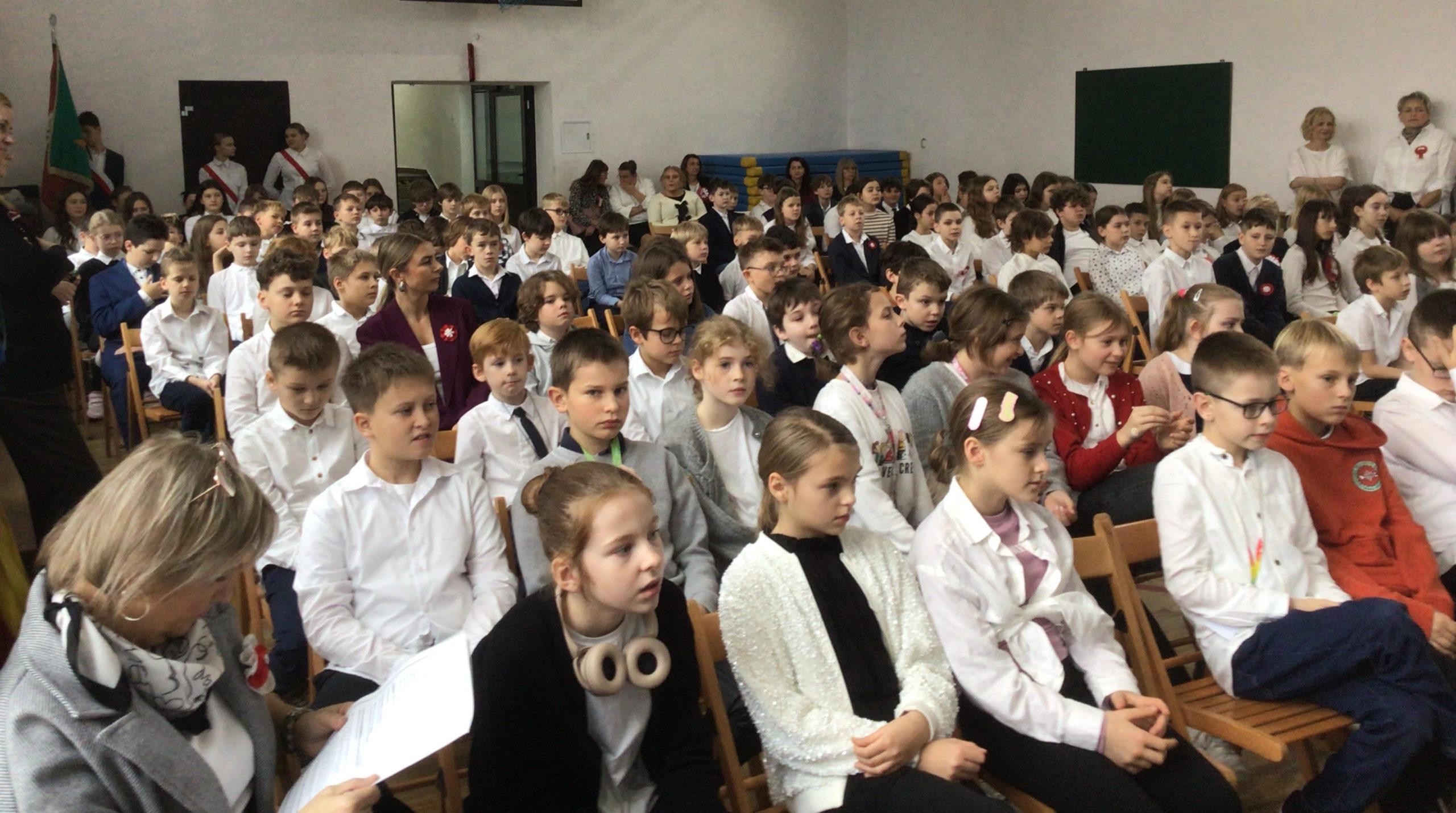 Na zdjęciu widać uczniów i nauczycieli zgromadzonych w sali gimnastycznej podczas szkolnej akademii z okazji Narodowego Święta Niepodległości. Uczestnicy siedzą na krzesłach, uważnie obserwując przebieg uroczystości. Większość uczniów ubrana jest odświętnie — w białe koszule, czasem z dodatkami w barwach narodowych. W tle widać sztandar szkolny oraz nauczycieli stojących przy ścianach sali.