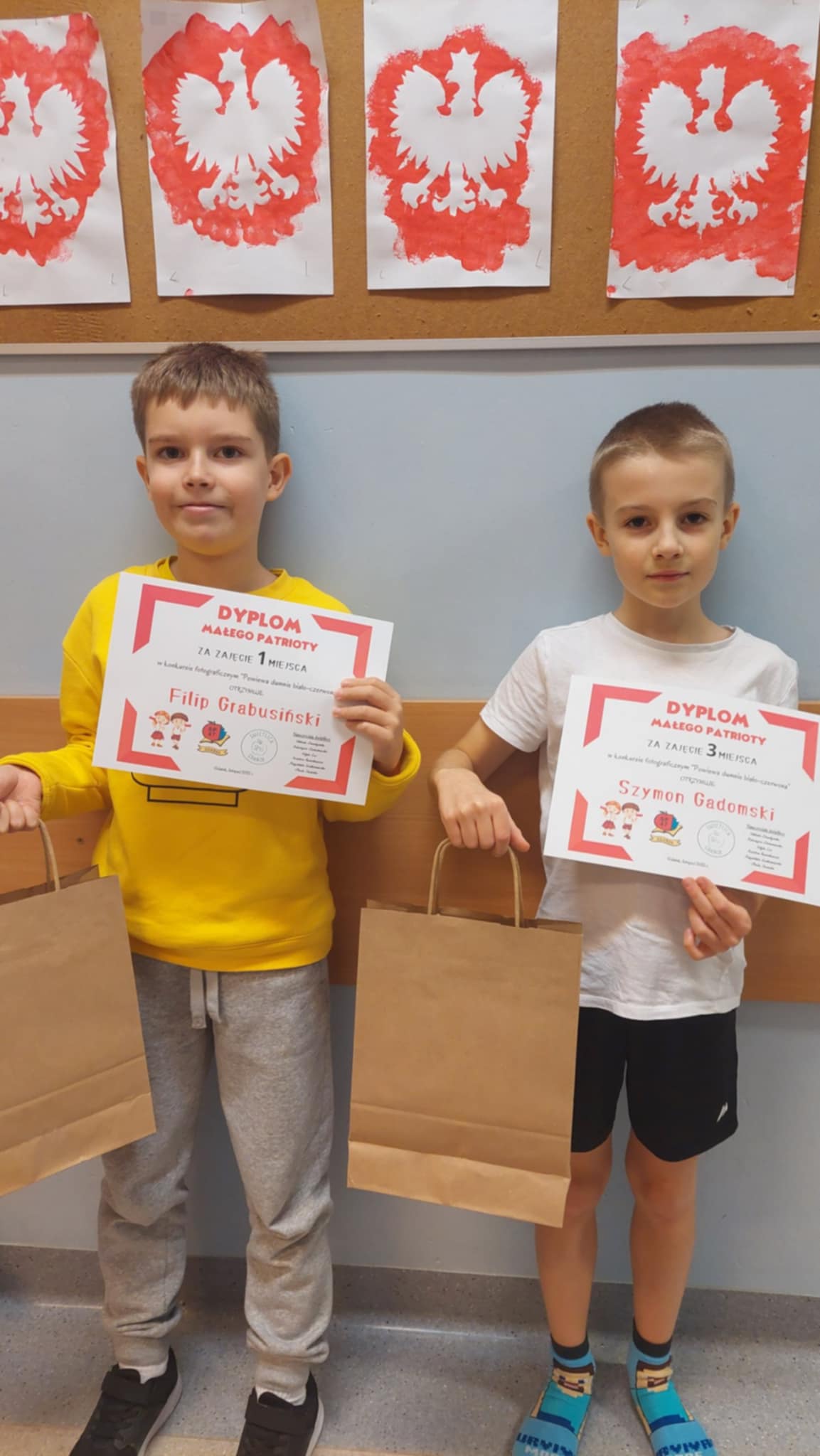 Dwóch chłopców stoi przy ścianie, każdy trzyma dyplom oraz papierową torbę z upominkiem. Za nimi widoczne są prace plastyczne z biało-czerwonym godłem.
