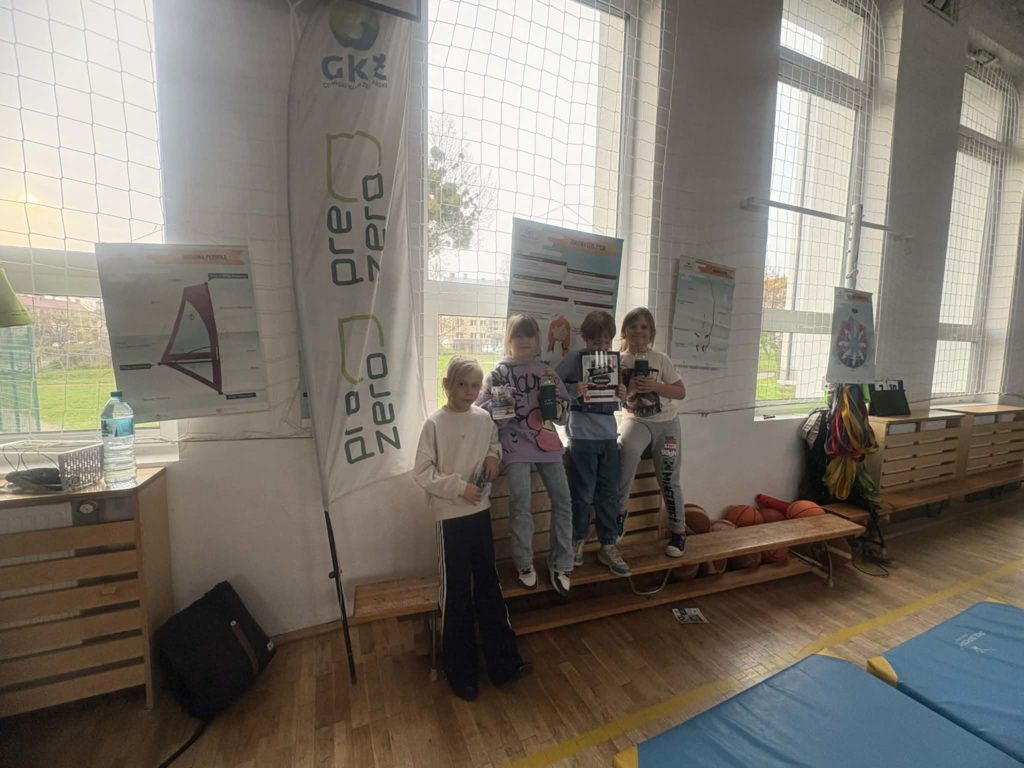 Czterech uczniów na sali gimnastycznej stoi przy oknie, w rękach trzymają prezenty od organizatora warsztatów