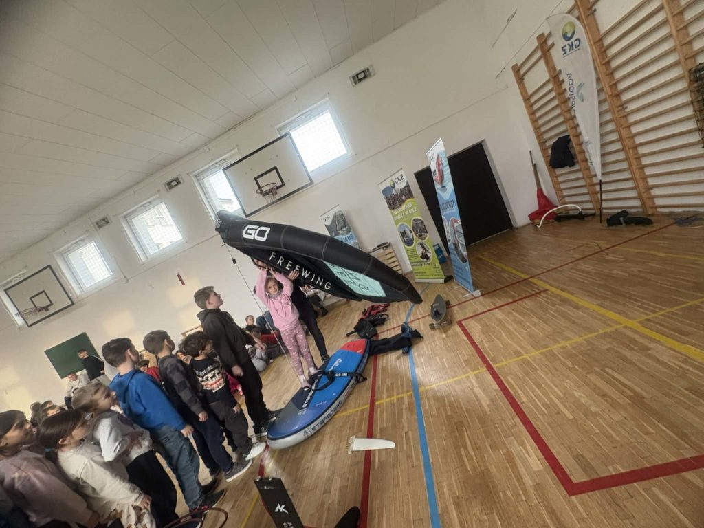 Na sali gimnastycznej jedno dziecko stoi na desce do pływania i trzyma dmuchany żagiel, grupka dzieci czeka na swoją kolej