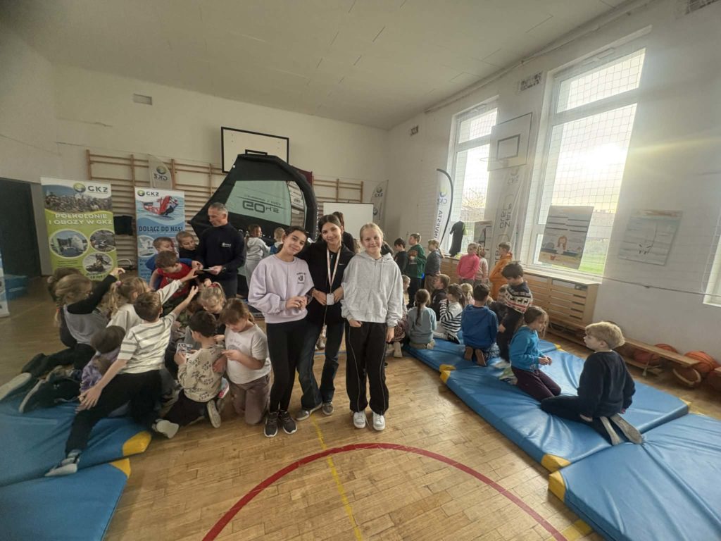 grupa uczniów siedzi na materacach, trzy dziewczynki stoją na tle żagla