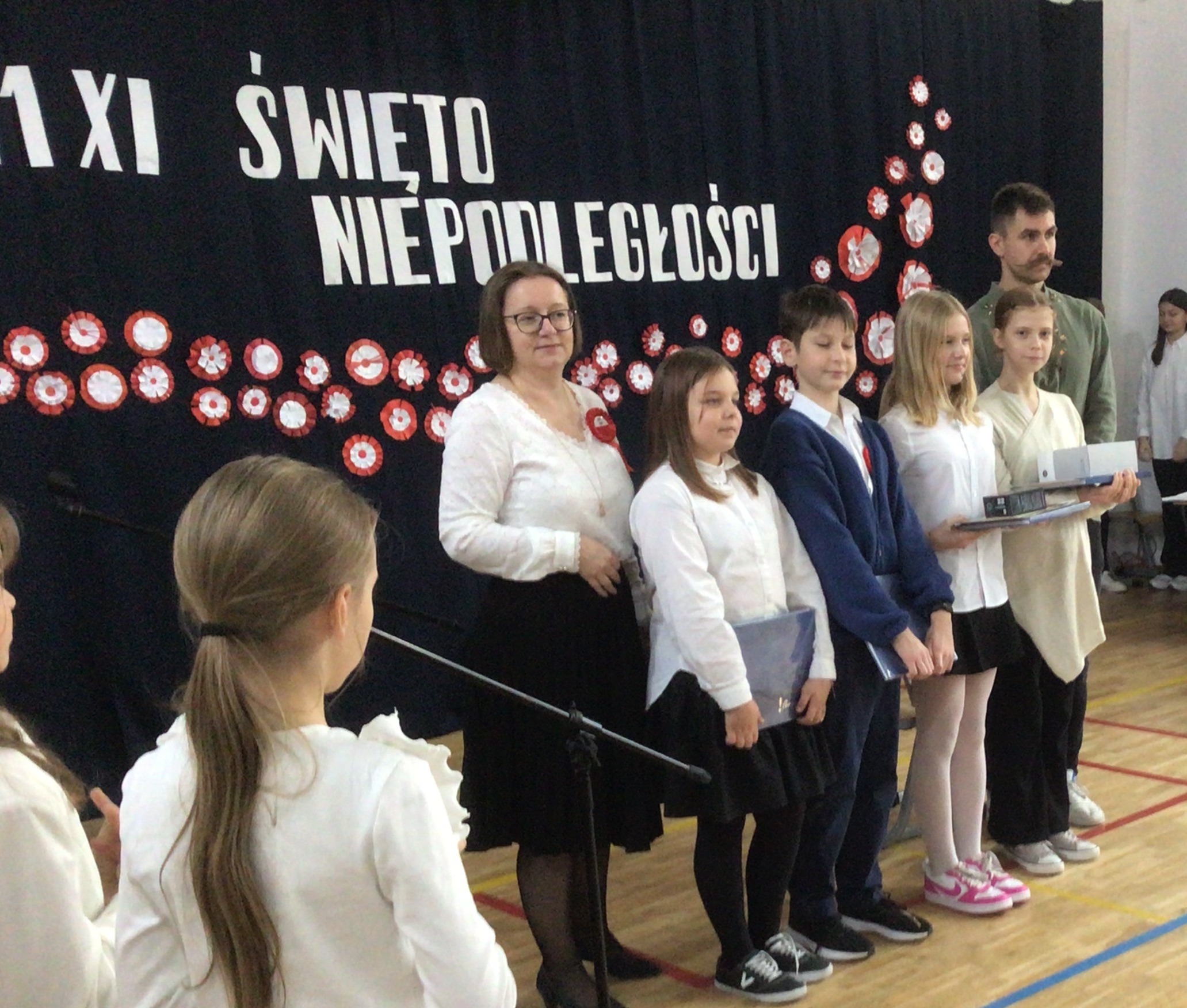 Na zdjęciu widać moment wręczenia nagród podczas szkolnej akademii z okazji Narodowego Święta Niepodległości. Na tle dekoracji z napisem „11 XI ŚWIĘTO NIEPODLEGŁOŚCI” stoi grupa uczniów w odświętnych strojach – białych koszulach i ciemnych spodniach lub spódnicach – trzymających nagrody. Obok nich znajdują się nauczyciele, którzy gratulują laureatom. Tło zdobią biało-czerwone kotyliony symbolizujące barwy narodowe.