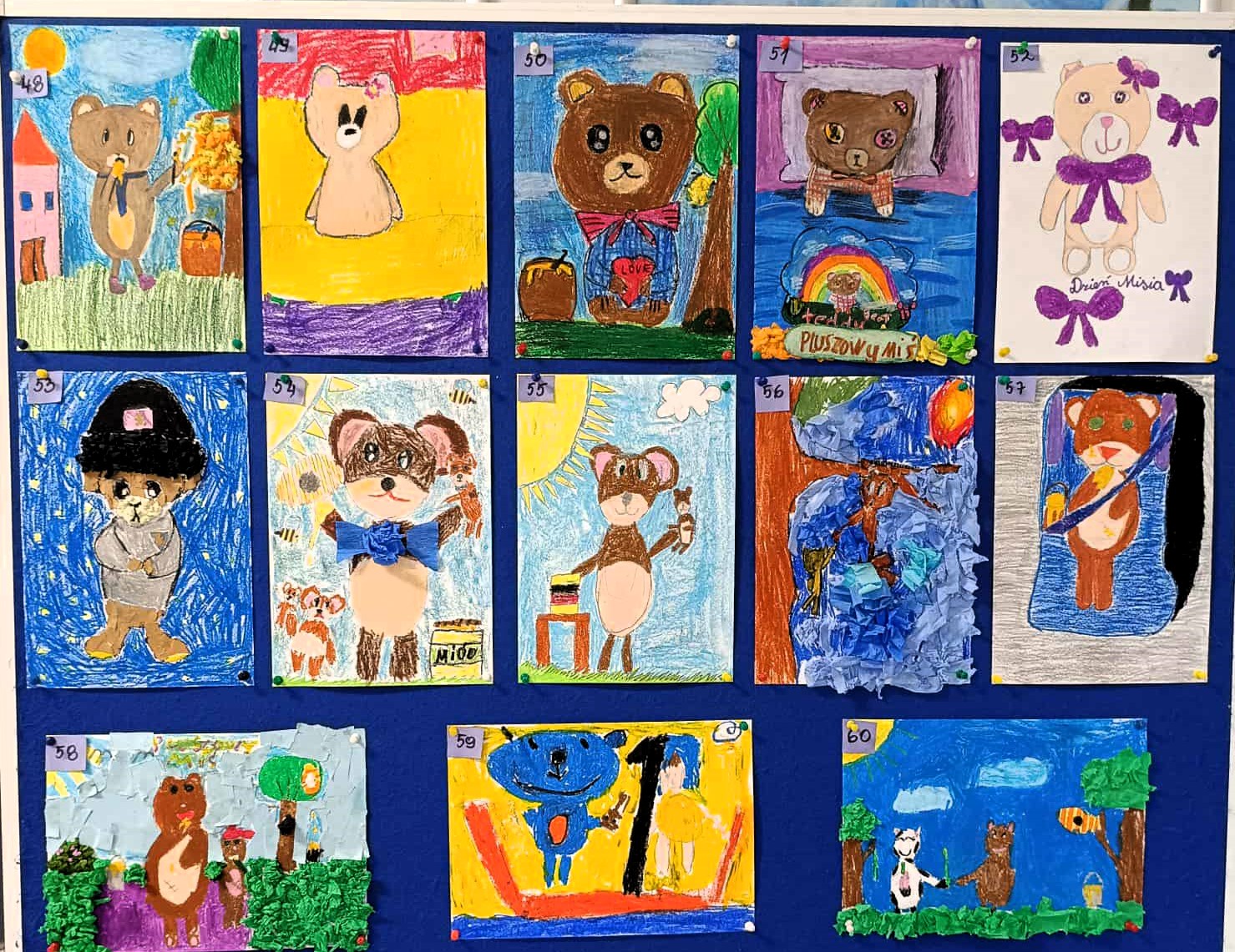 Zdjęcie przedstawia tablicę ekspozycyjną z kolorowymi pracami plastycznymi dzieci, uczestników konkursu „Świetlicowy Konkurs Plastyczny – Miś”. Na ciemnoniebieskim tle przypiętych jest kilkanaście rysunków misiów wykonanych różnymi technikami. 