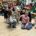 Na środku sali siedzi biały pies Kajtek dookoła niego siedzą dzieci