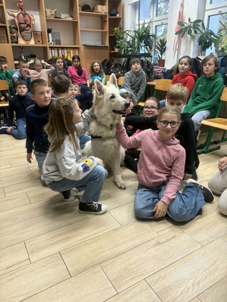 Na środku sali siedzi biały pies Kajtek dookoła niego siedzą dzieci