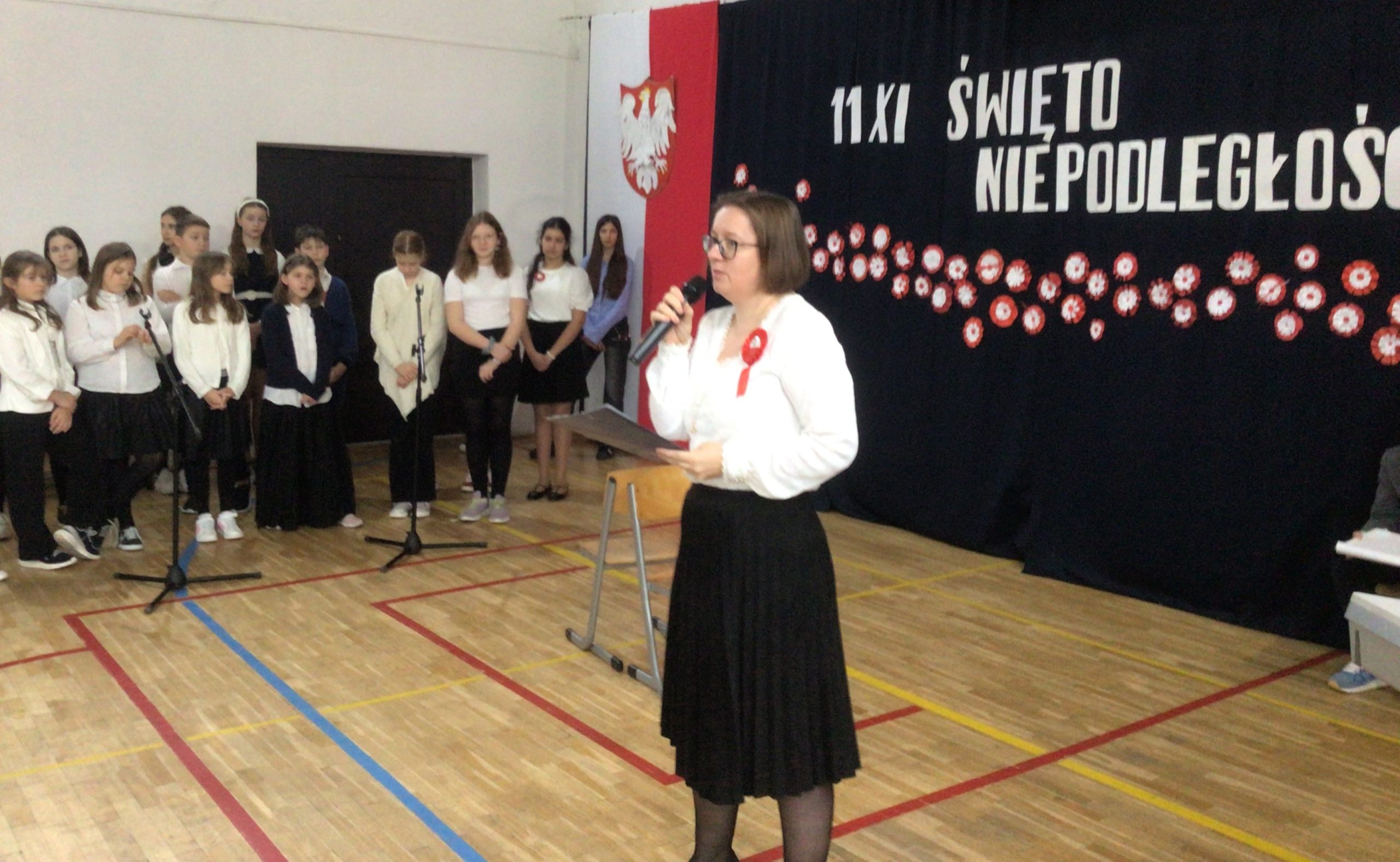 Na zdjęciu widać szkolną akademię z okazji Narodowego Święta Niepodległości obchodzonego 11 listopada. Na tle ciemnej kurtyny z napisem „11 XI ŚWIĘTO NIEPODLEGŁOŚCI” oraz dekoracją z biało-czerwonych rozet stoi kobieta w białej bluzce i czarnej spódnicy, przemawiająca do mikrofonu. Na jej piersi przypięty jest biało-czerwony kotylion. W tle stoi grupa uczniów ubranych w galowe stroje – białe koszule i ciemne spódnice lub spodnie. Scena odbywa się w sali gimnastycznej, udekorowanej w barwach narodowych Polski.