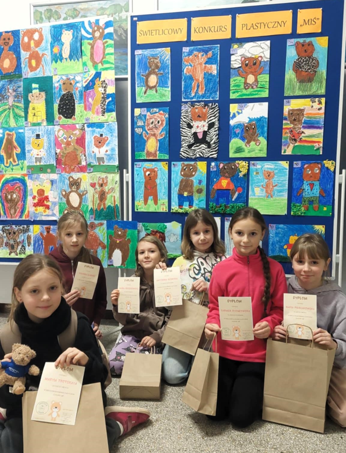 Grupa sześciu uczennic siedzi na podłodze przed wystawą kolorowych prac plastycznych przedstawiających misie. Dziewczynki trzymają dyplomy i papierowe torebki z nagrodami. W tle widać dwie duże tablice z pracami konkursowymi.