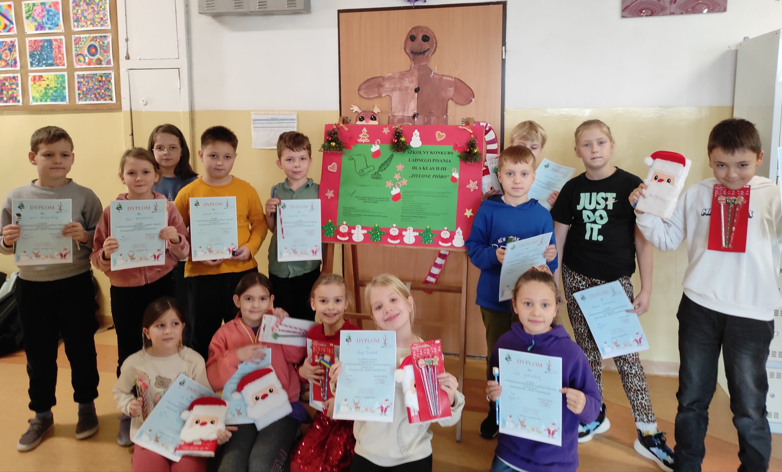 Uczestnicy konkursu ustawieni w dwa rzędy, pierwszy kuca, drugi stoi. Dzieci trzymają w dłoniach dyplomy i nagrody. W tle tablica z informacjami dotyczącymi konkursu kaligraficznego, drzwi, do których przyczepione są ozdoby świąteczne oraz ściana.