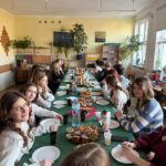 Grupa uczniów z klasy 8b siedzą przy długim stole nakrytym zielonym obrusem w sali lekcyjnej, na którym znajdują się talerze, napoje i świąteczne przekąski, podczas wspólnego klasowego spo-tkania.