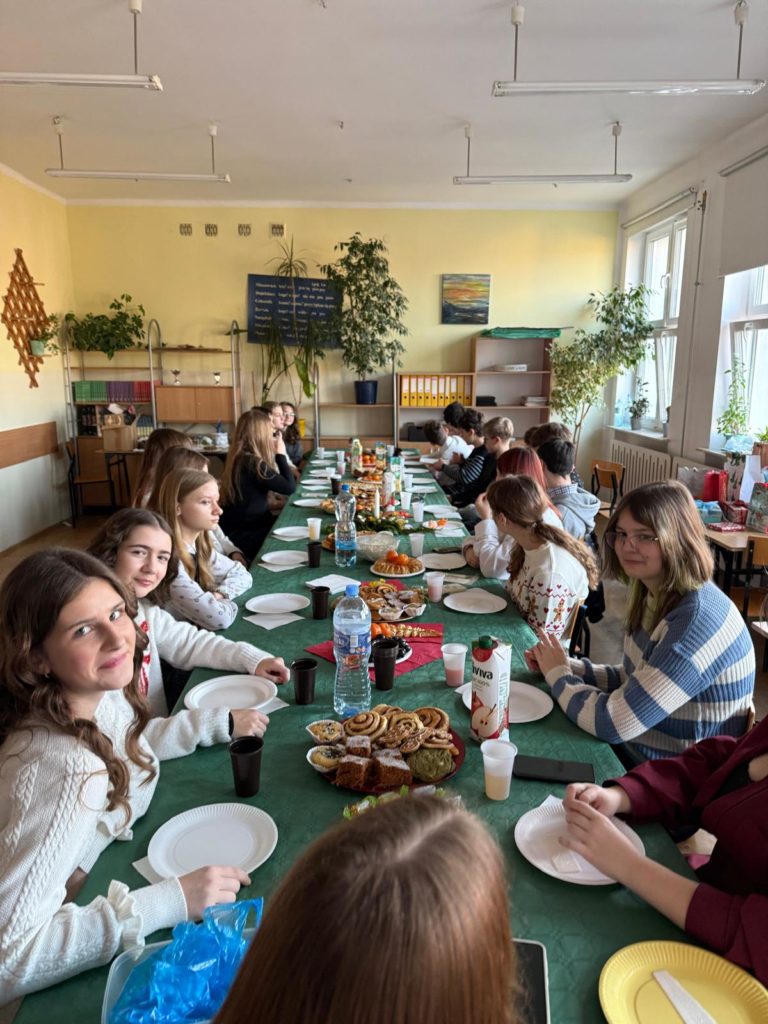 Grupa uczniów z klasy 8b siedzą przy długim stole nakrytym zielonym obrusem w sali lekcyjnej, na którym znajdują się talerze, napoje i świąteczne przekąski, podczas wspólnego klasowego spo-tkania.