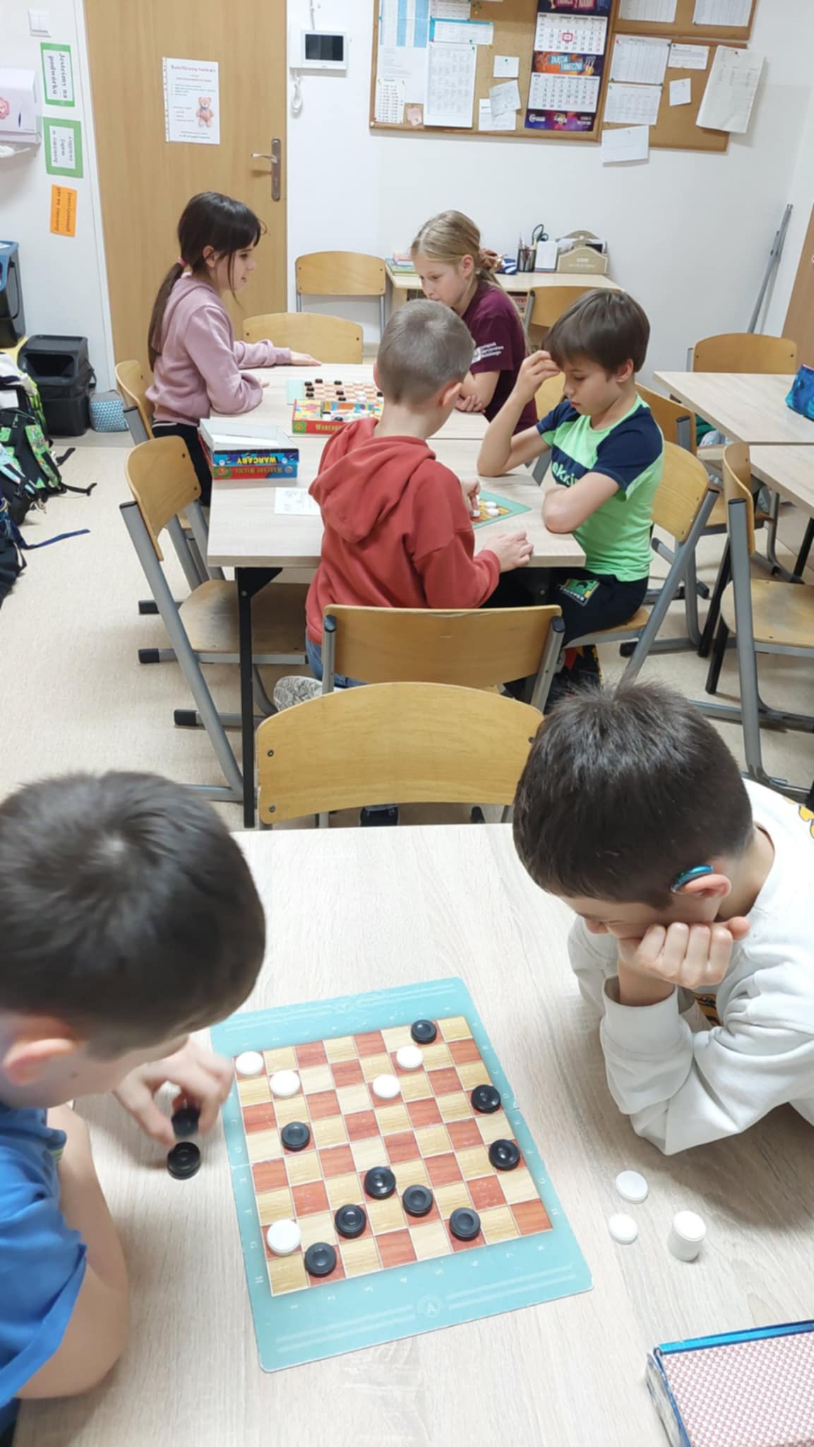 Kilka grup dzieci siedzi przy stolikach w szkolnej świetlicy i gra w warcaby. Widać skupionych uczestników przy planszach, pochylonych nad pionkami.