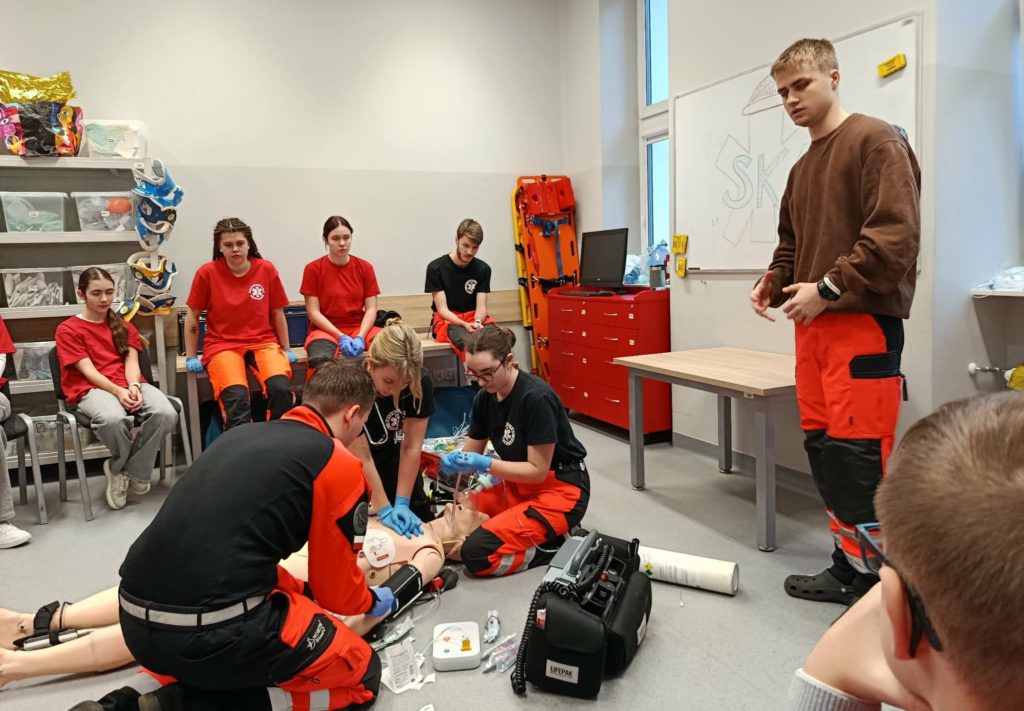 Na pierwszym planie troje studentów ratownictwa udzielających pomocy poszkodowanemu podczas pozoracji, obok nich inny stojący student i monitor do badania parametrów życiowych. W tle uczennica koła HOPR i inni przyglądający się studenci.