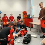 Na pierwszym planie troje studentów ratownictwa udzielających pomocy poszkodowanemu podczas pozoracji, obok nich inny stojący student i monitor do badania parametrów życiowych. W tle uczennica koła HOPR i inni przyglądający się studenci.