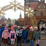 Na tle gdańskich kamienic pod łukiem iluminacji ze śnieżynkami stoi grupa uczniów klasy 1c. Po bokach stragany jarmarku.