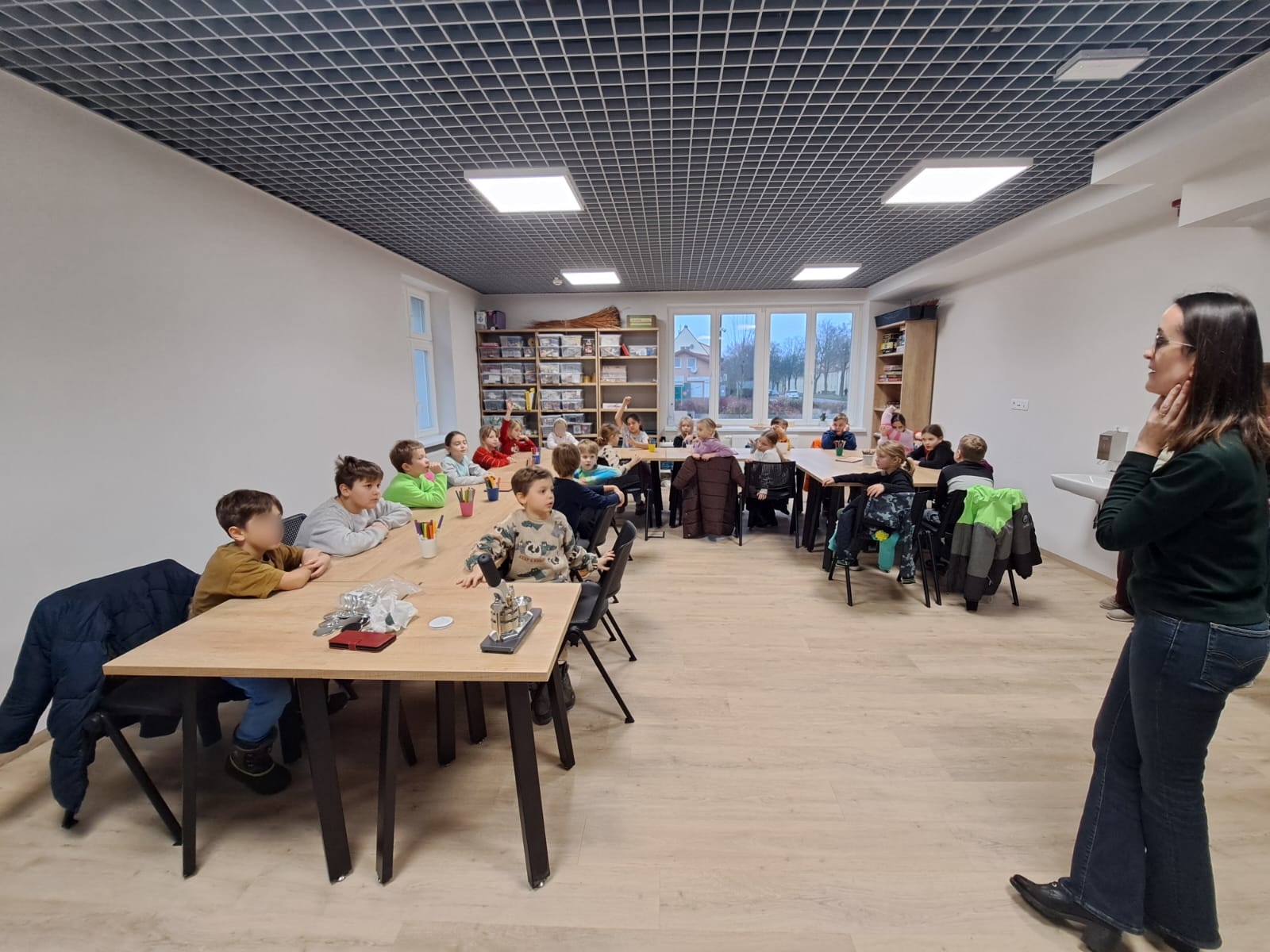 Grupa dzieci siedzi przy stołach w jasnej sali warsztatowej, słuchając prowadzącej stojącej z prawej strony.