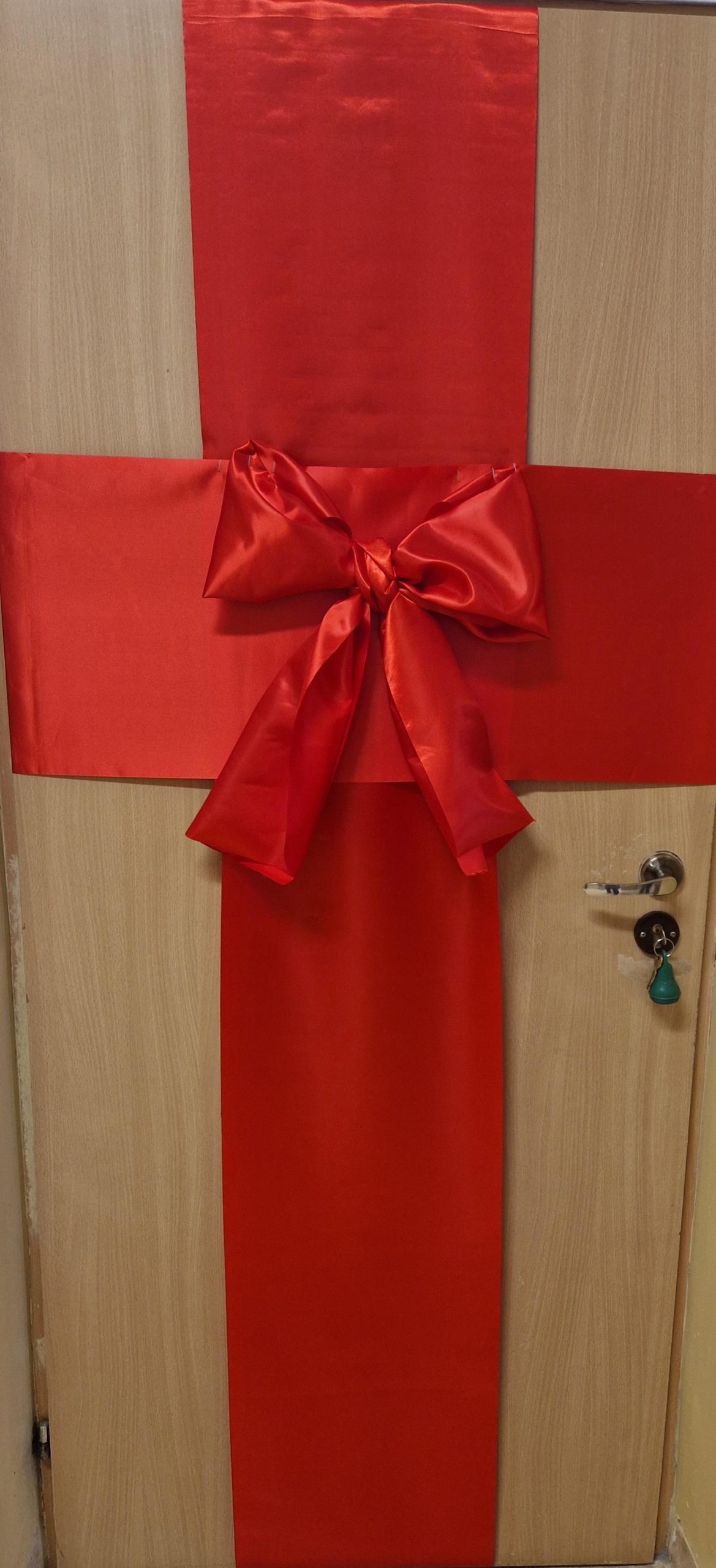 Drewniane drzwi udekorowane jak prezent: przez środek pionowo i poziomo biegną szerokie czerwone wstęgi tworzące krzyż. Na środku znajduje się duża czerwona kokarda. Po prawej stronie widoczna klamka z kluczem, drzwi osadzone w jasnej futrynie.