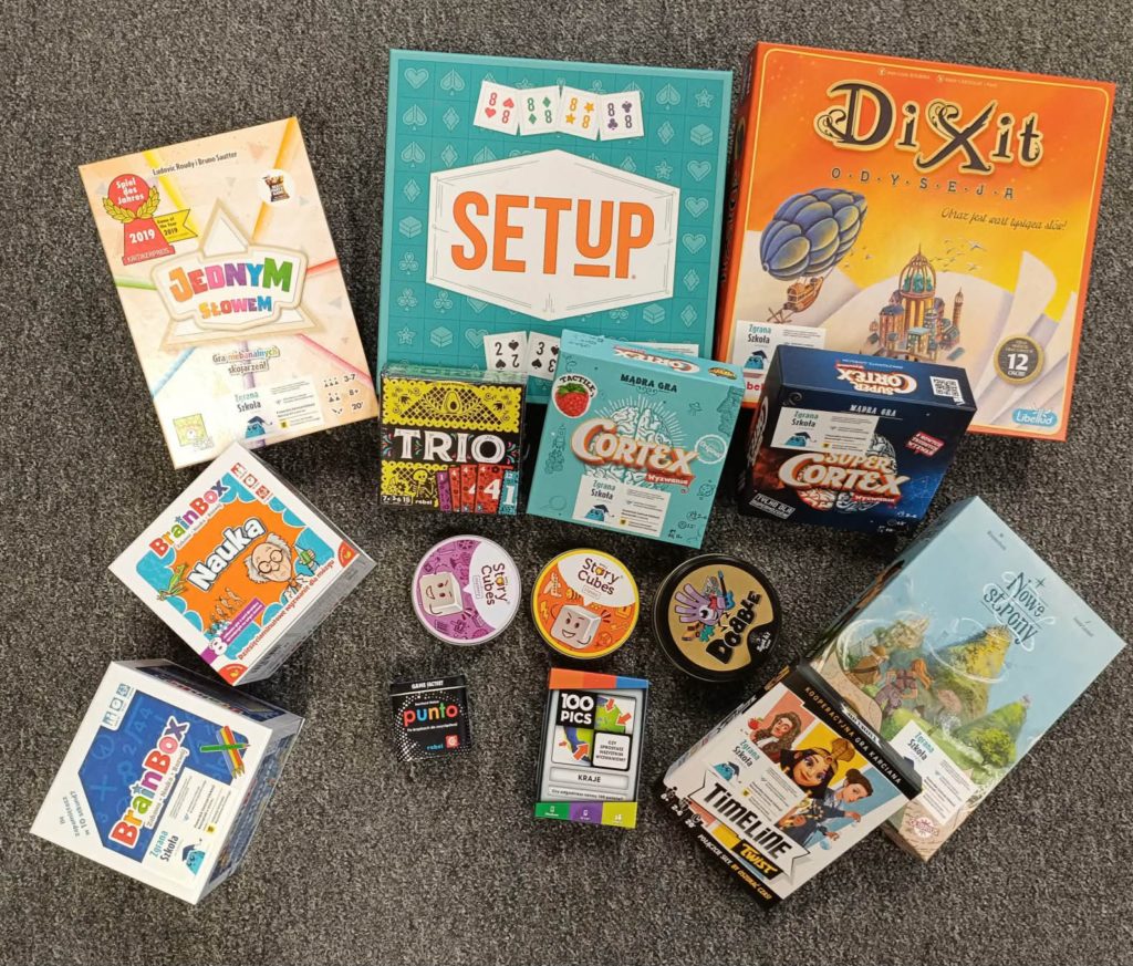 : Zdjęcie przedstawia kilkanaście pudełek gier planszowych i karcianych rozłożonych na szarej wykładzinie. Wśród nich znajdują się m.in. „Setup”, „Dixit Odyseja”, „Jednym Słowem”, „Cortex” i „Super Cortex”, „Dobble”, „Story Cubes”, „Trio”, „Punto”, „100 Pics”, „Timeline Twist”, „BrainBox Nauka” oraz inne małe gry familijne. Wszystkie pudełka są kolorowe i ułożone w lekkim nieładzie.