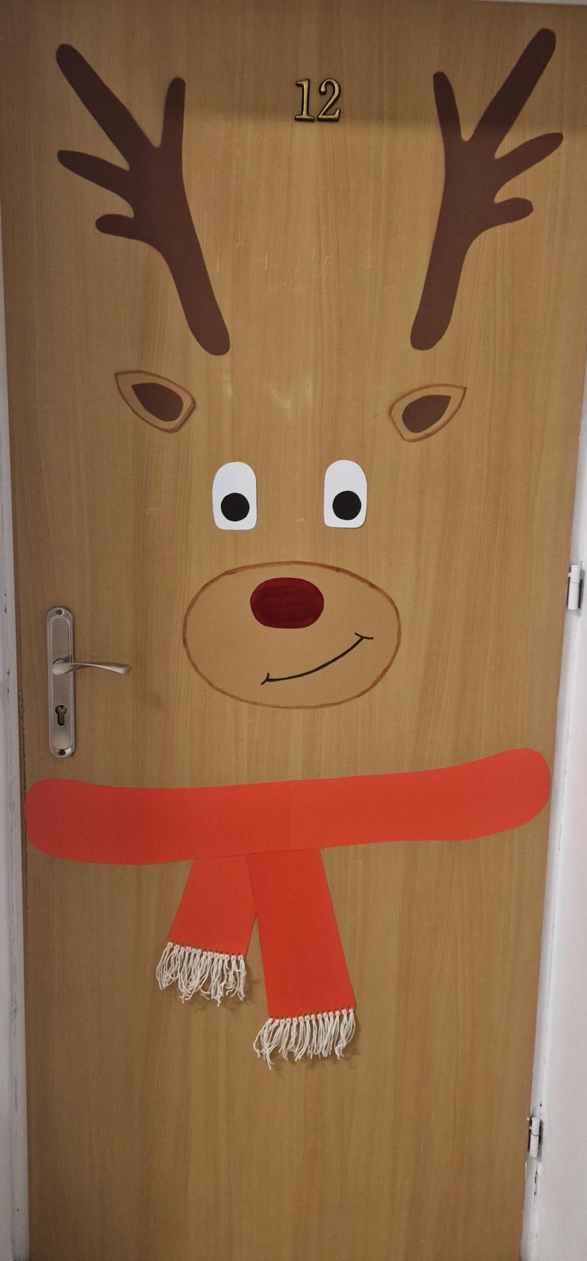 Drewniane drzwi ozdobione jak renifer: na górze numer „12”, narysowane brązowe poroże i uszy, duże oczy, czerwony nos i uśmiech. Na dole drzwi wisi czerwona szalikowa ozdoba z frędzlami. Po lewej stronie klamka, drzwi w białej futrynie.