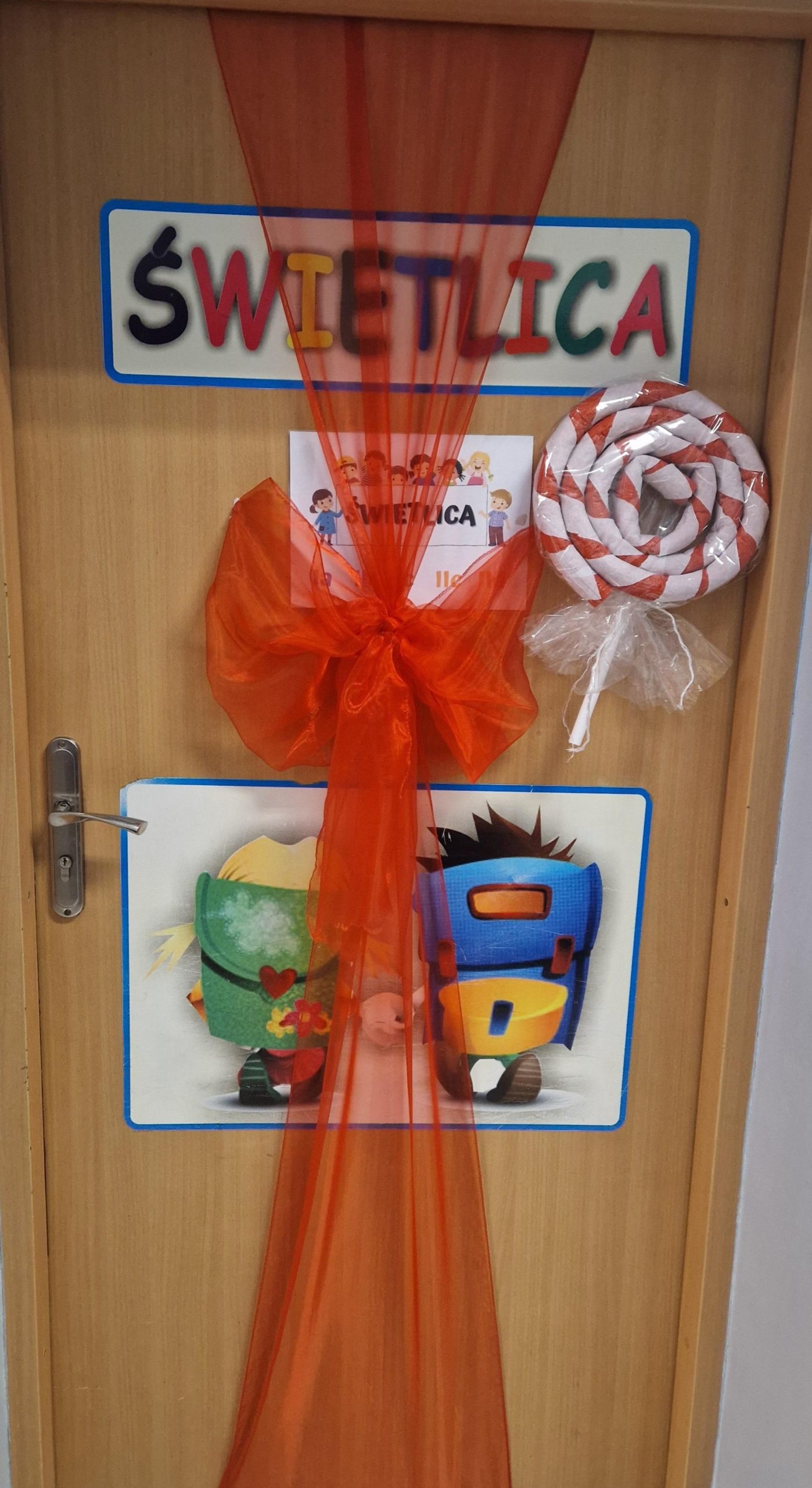 Drewniane drzwi ozdobione dekoracjami świetlicy: u góry kolorowy napis „ŚWIETLICA”. Przez środek drzwi biegnie półprzezroczysta czerwona wstęga z dużą kokardą. Po prawej stronie znajduje się ozdobny lizak w biało-czerwone paski. Na dole umieszczona jest ilustracja dzieci z plecakami. Po lewej stronie widoczna klamka, drzwi osadzone w jasnej futrynie.