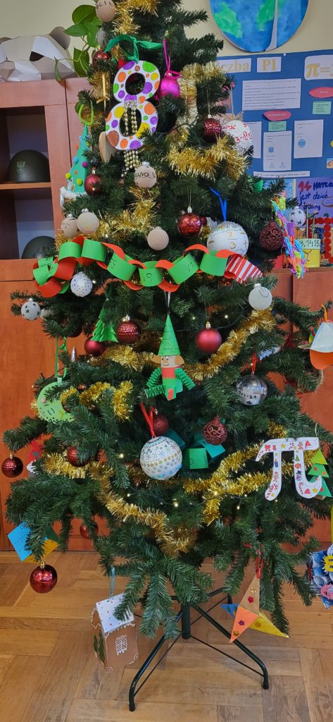 Szkolna dekoracja świąteczna w matematycznym stylu. Choinka i lampiony są ozdobione własnoręcznie wykonanymi ozdobami z figurami geometrycznymi, liczbami i symbolem π. Całość tworzy kolorową i pomysłową ekspozycję przygotowaną na szkolny konkurs.