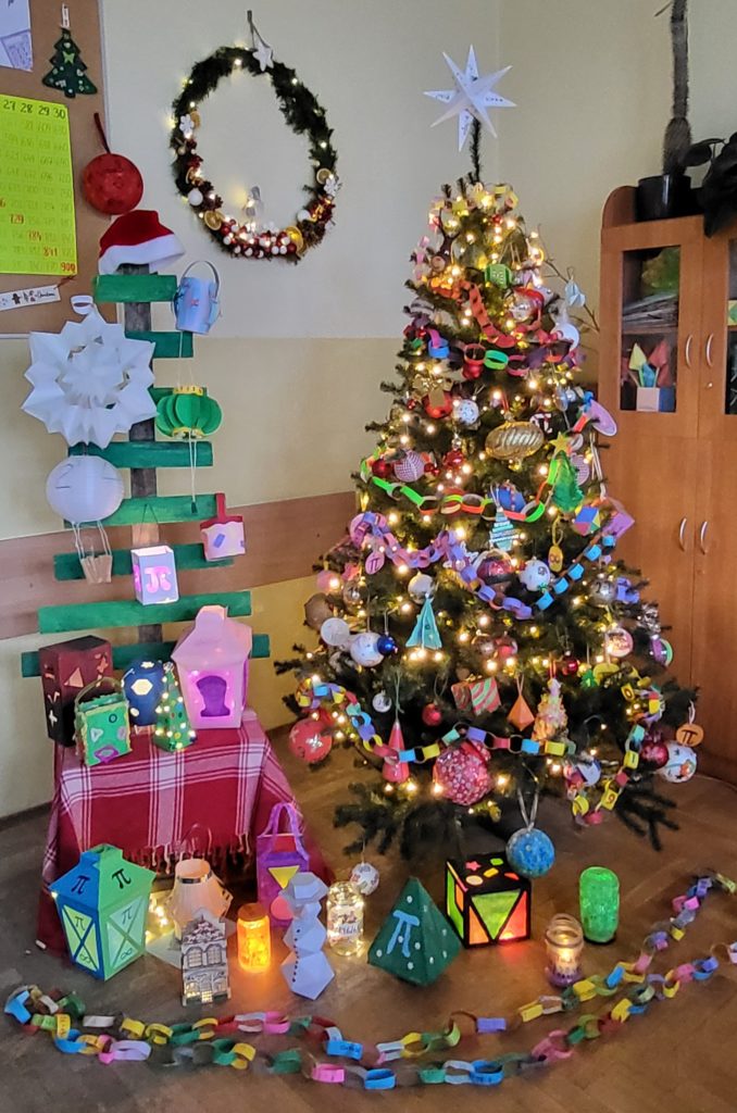 Szkolna dekoracja świąteczna w matematycznym stylu. Choinka i lampiony są ozdobione własnoręcznie wykonanymi ozdobami z figurami geometrycznymi, liczbami i symbolem π. Całość tworzy kolorową i pomysłową ekspozycję przygotowaną na szkolny konkurs.