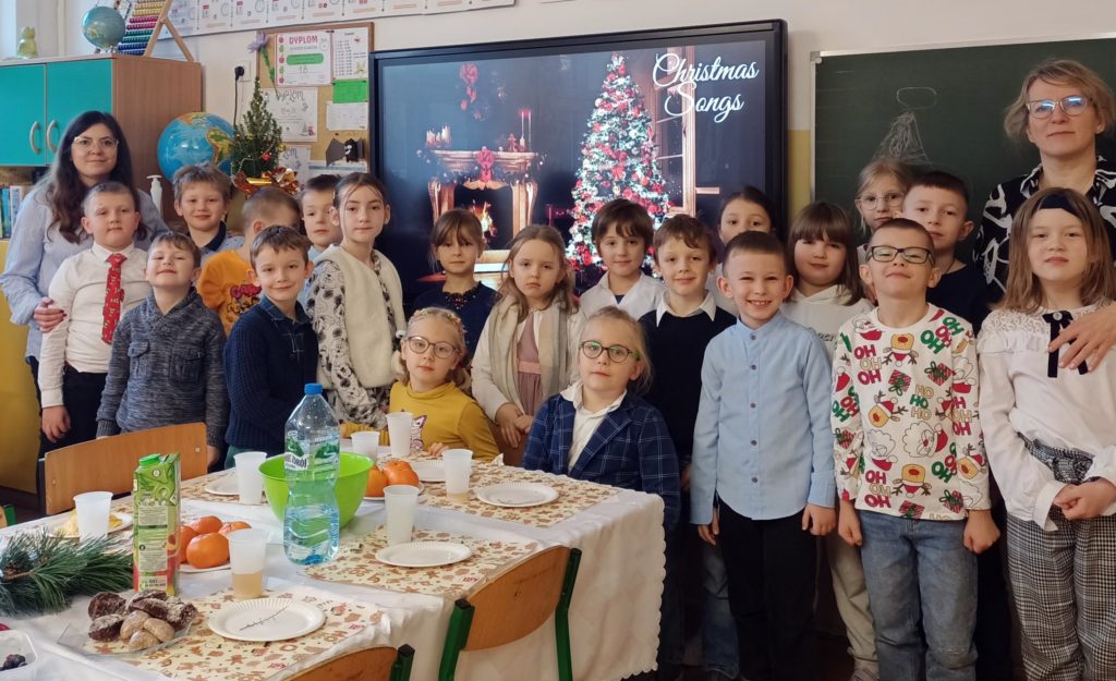 Uczniowie ustawieni w dwóch rzędach na tle ekranu, na którym wyświetla się dekoracja świąteczna z choinką. Po prawej i lewej stronie stoją nauczycielki. Przed nim dwoje dzieci siedzących przy świątecznym stole nakrytym białym obrusem. Na stole leżą talerzyki, ciastka i sosnowe gałązki. 
