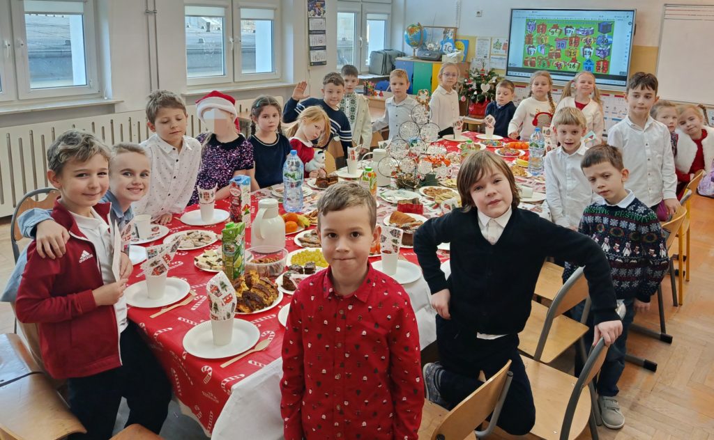 W koło zastawionego stołu wigilijnego stoi dwudziestoosobowa grupa uczniów klasy 1c ubrana w świąteczne stroje pozując do zdjęcia. W tle tablica multimedialna z kalendarzem adwentowym.