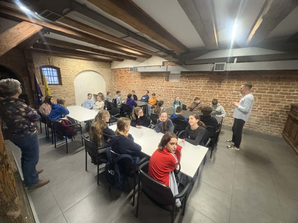 Uczniowie siedzą przy stołach i słuchają przewodnika