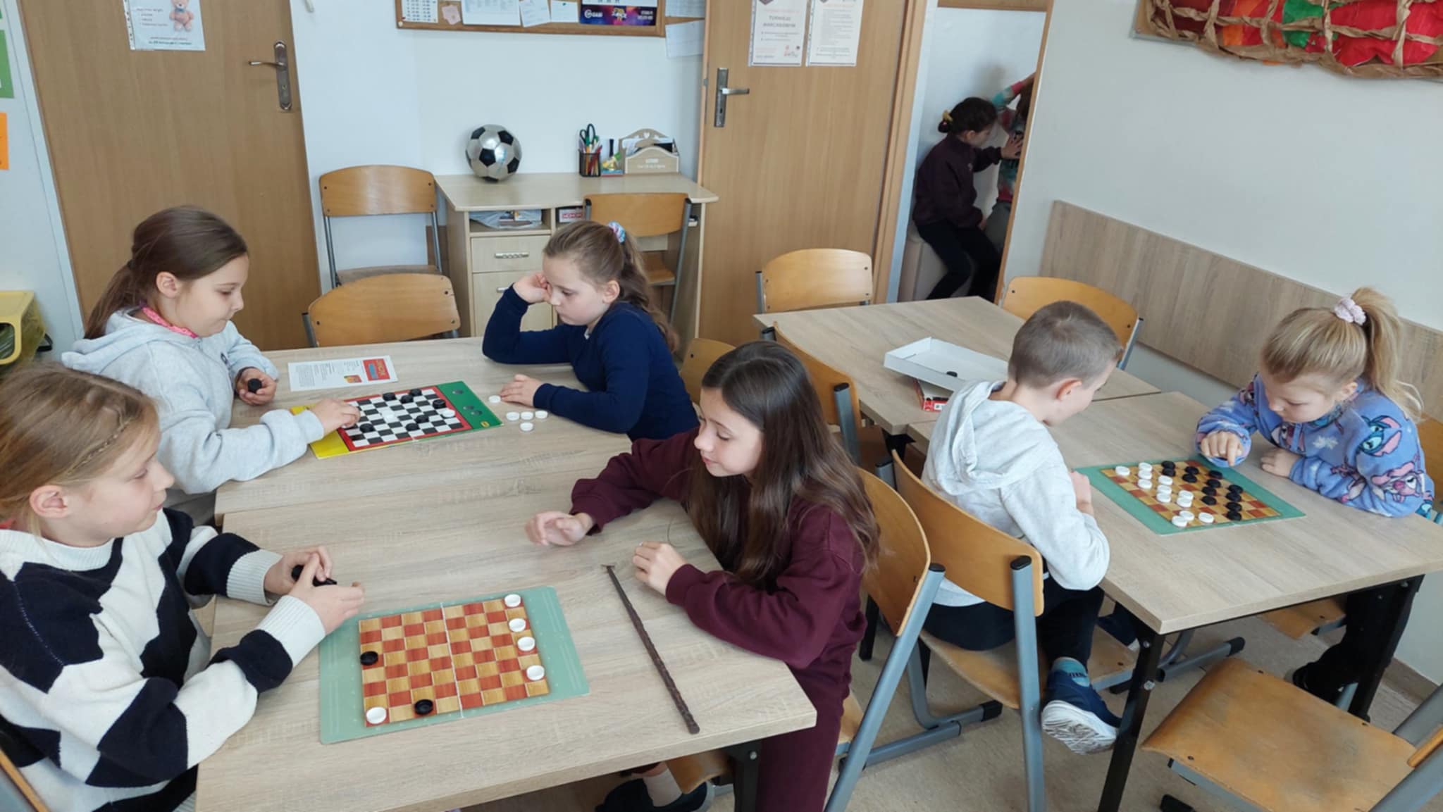 Dzieci siedzą przy stołach w świetlicy i rozgrywają partie warcabów. Każda para gra na swojej planszy, a atmosfera jest pełna skupienia.