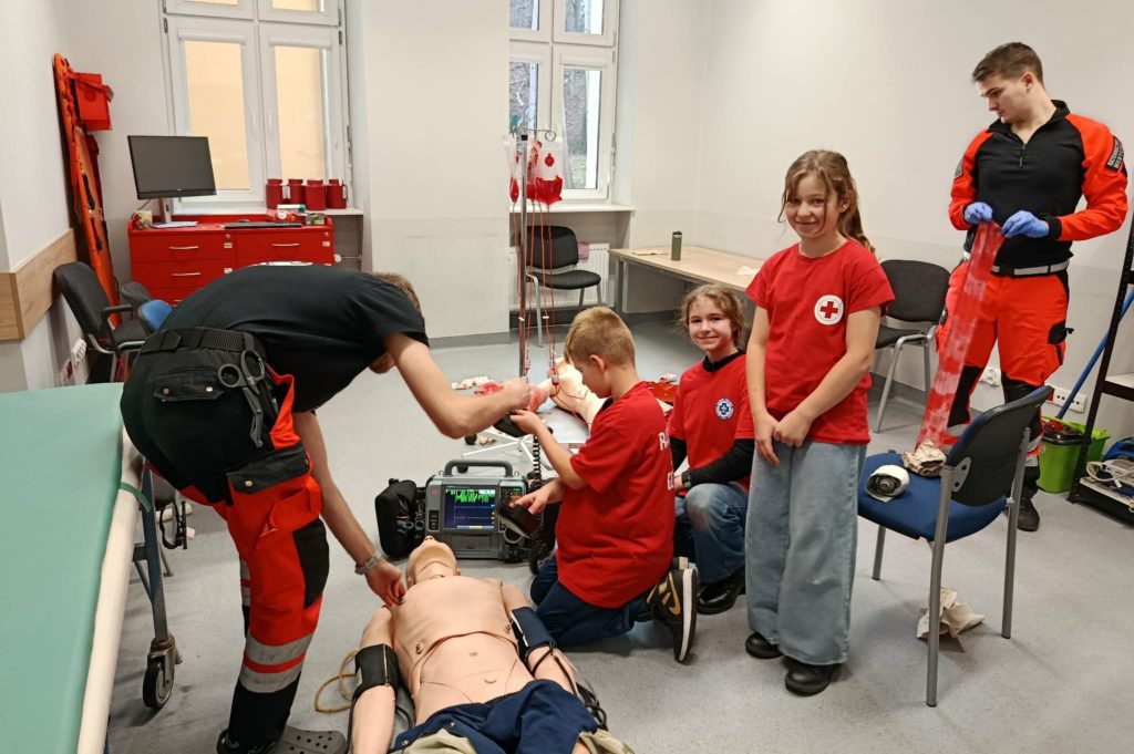 Na pierwszym planie leżący na podłodze fantom, monitor z elektrodami, obok trzech uczniów Koła PP HOPR obsługujących sprzęt i dwaj studenci ratownictwa przyglądających się uczniom w akcji.