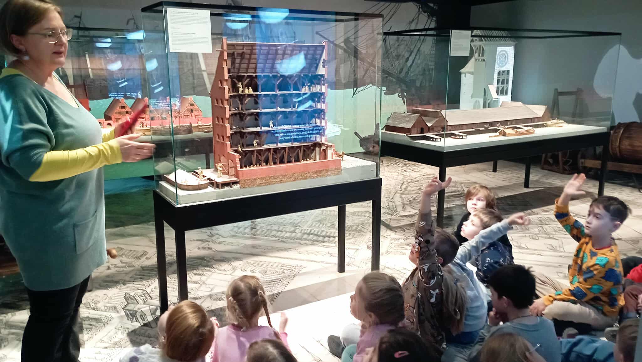 Na pierwszym planie kustoszka Żurawia stojąca przy szklanej gablocie z makietą spichlerza. Obok gablota z makietą nabrzeża portowego. Przed gablotą uczniowie klasy 1c
