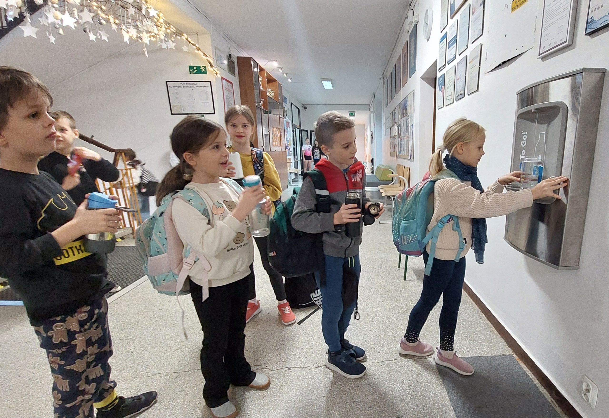 Grupa dzieci w szkolnym korytarzu stoi przy dystrybutorze wody. Jedna dziewczynka napełnia swoją butelkę, a pozostałe dzieci czekają w kolejce, trzymając własne bidony. Wszystkie mają plecaki i wyglądają.
