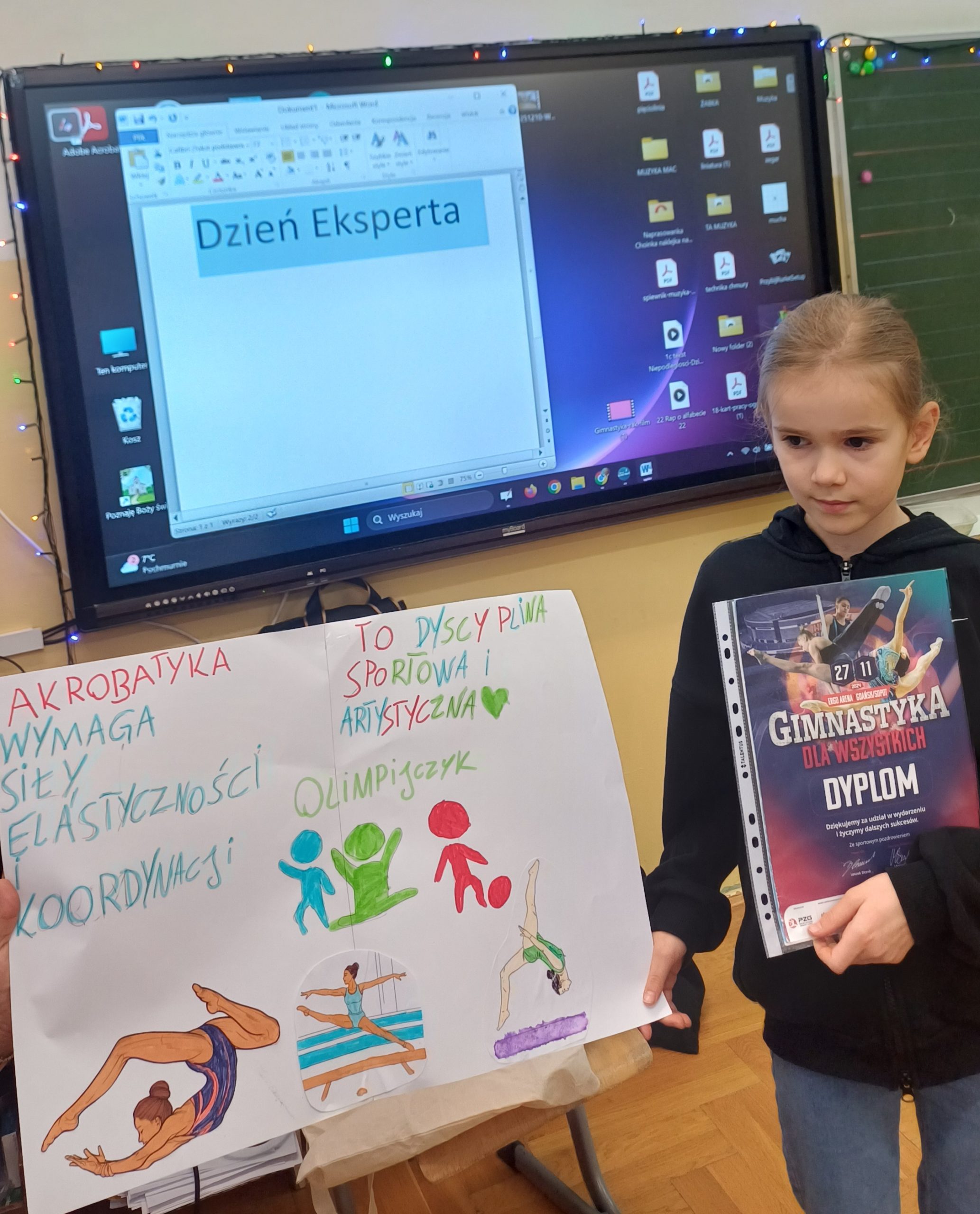 Dziewczynka stoi trzymając w jednej ręce dyplom, a w drugiej plakat o akrobatyce.
